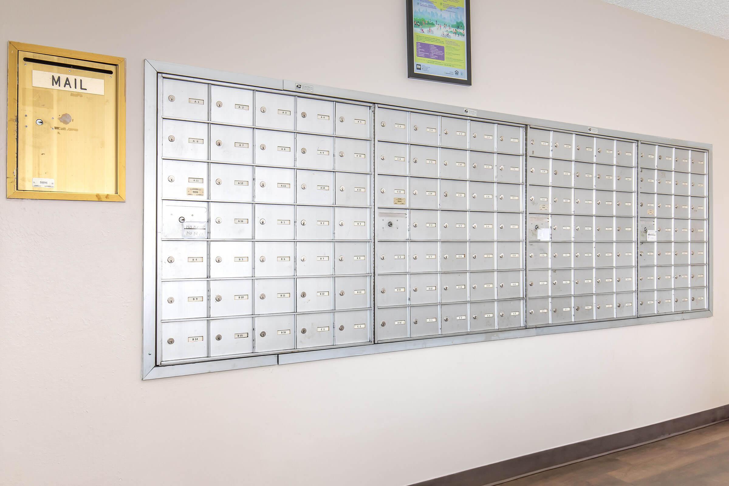 A wall-mounted postal unit featuring multiple metal mailboxes arranged in rows, with a wooden mailbox on the left side labeled "MAIL." The unit is set against a light-colored wall, and there is a framed picture or notice above the mailboxes.