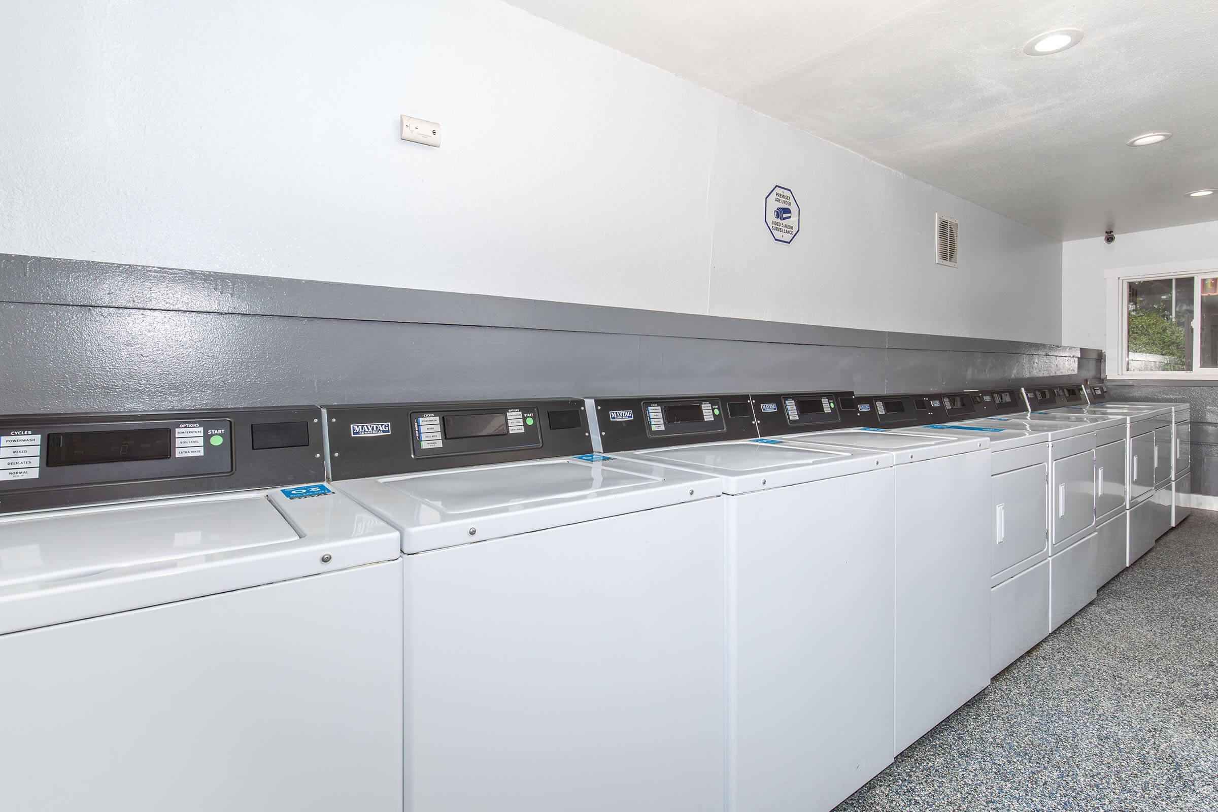 A clean, well-lit laundry room featuring several white washing machines and dryers lined up against a gray wall. The floor is made of gray tiles, and there is a ceiling light illuminating the space. A wall-mounted sign is visible in the background.