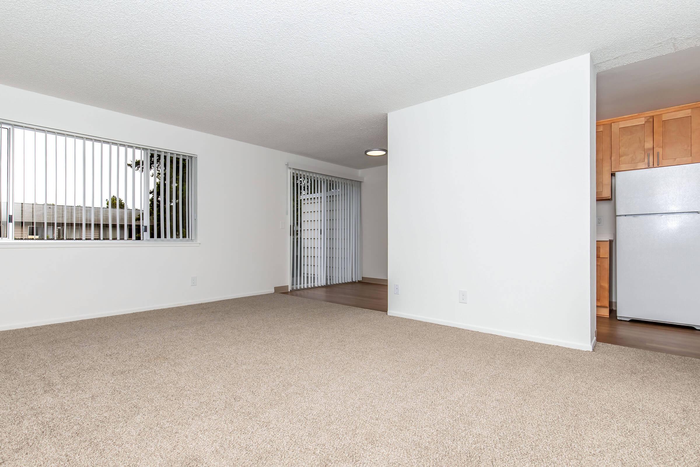 Empty living room with light beige carpet, white walls, and large window with vertical blinds. In the background, a sliding glass door leads to an outdoor area. A small kitchenette is visible to the right, featuring a white refrigerator. The space appears bright and newly renovated.