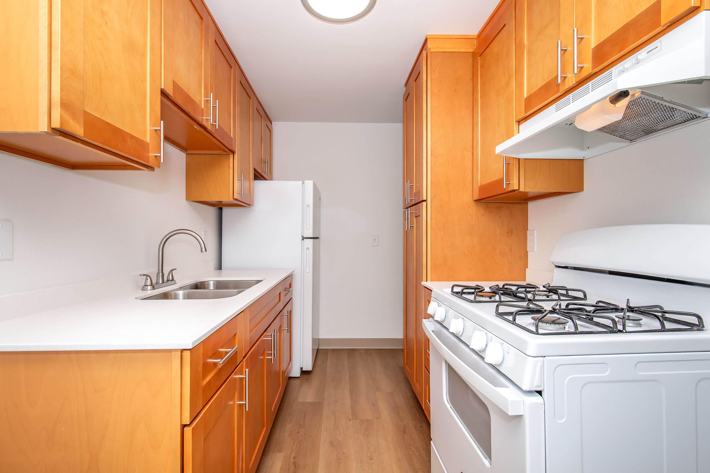 A modern kitchen featuring light wood cabinets, a white sink, and a gas stove. The kitchen includes a refrigerator and a clean countertop, with a well-lit and spacious layout, showcasing an organized interior design.