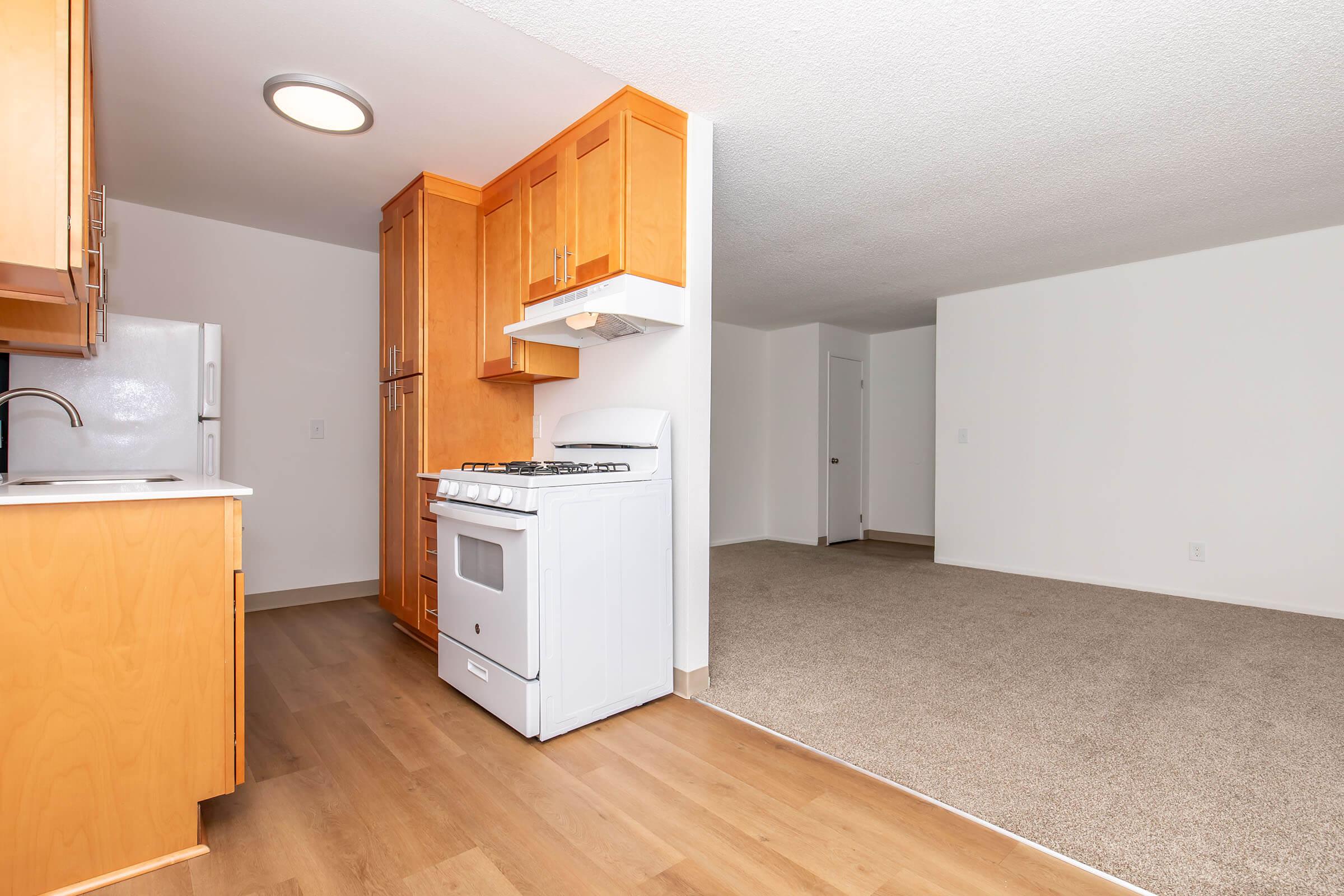 A kitchen area featuring wooden cabinets, a gas stove, and a refrigerator. In the background, there is a spacious living area with carpeted flooring and white walls, leading to an open doorway. The layout creates a feeling of openness between the kitchen and living space.