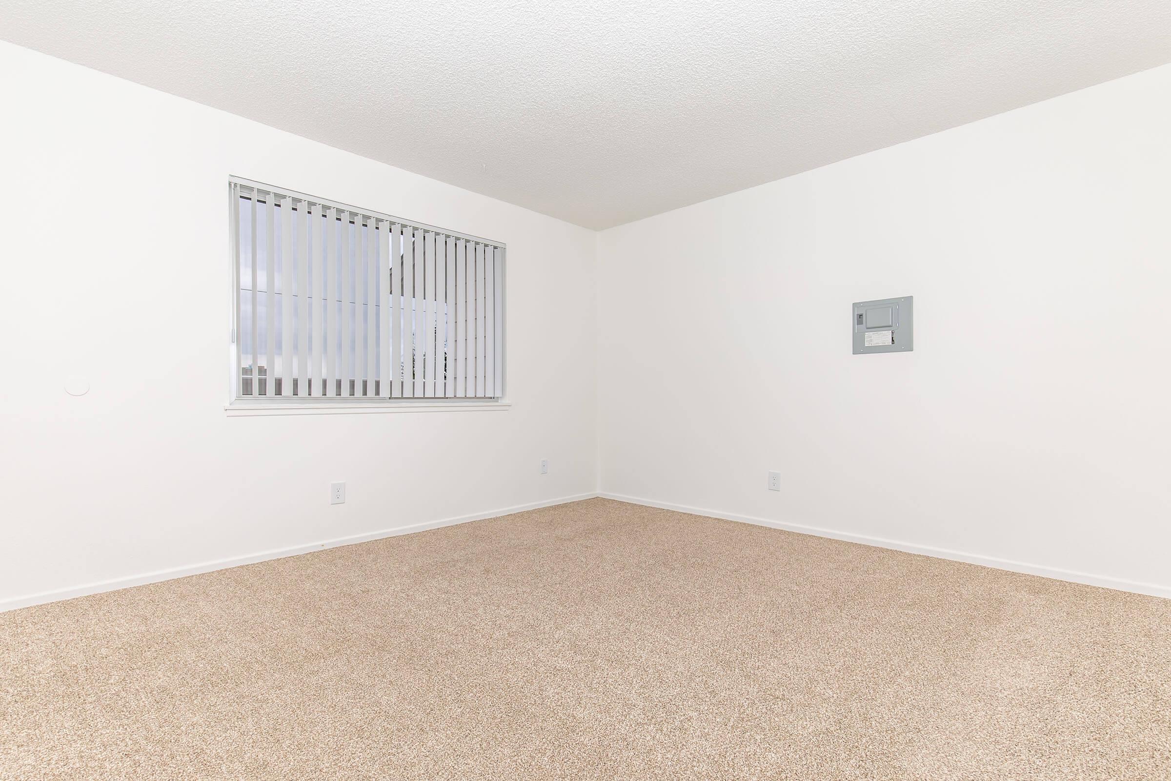 A spacious, empty room with beige carpeting and white walls. A single window with vertical blinds allows natural light to brighten the space. There is a small electrical panel mounted on the wall. The overall ambiance is clean and minimalist, ready for personalization.