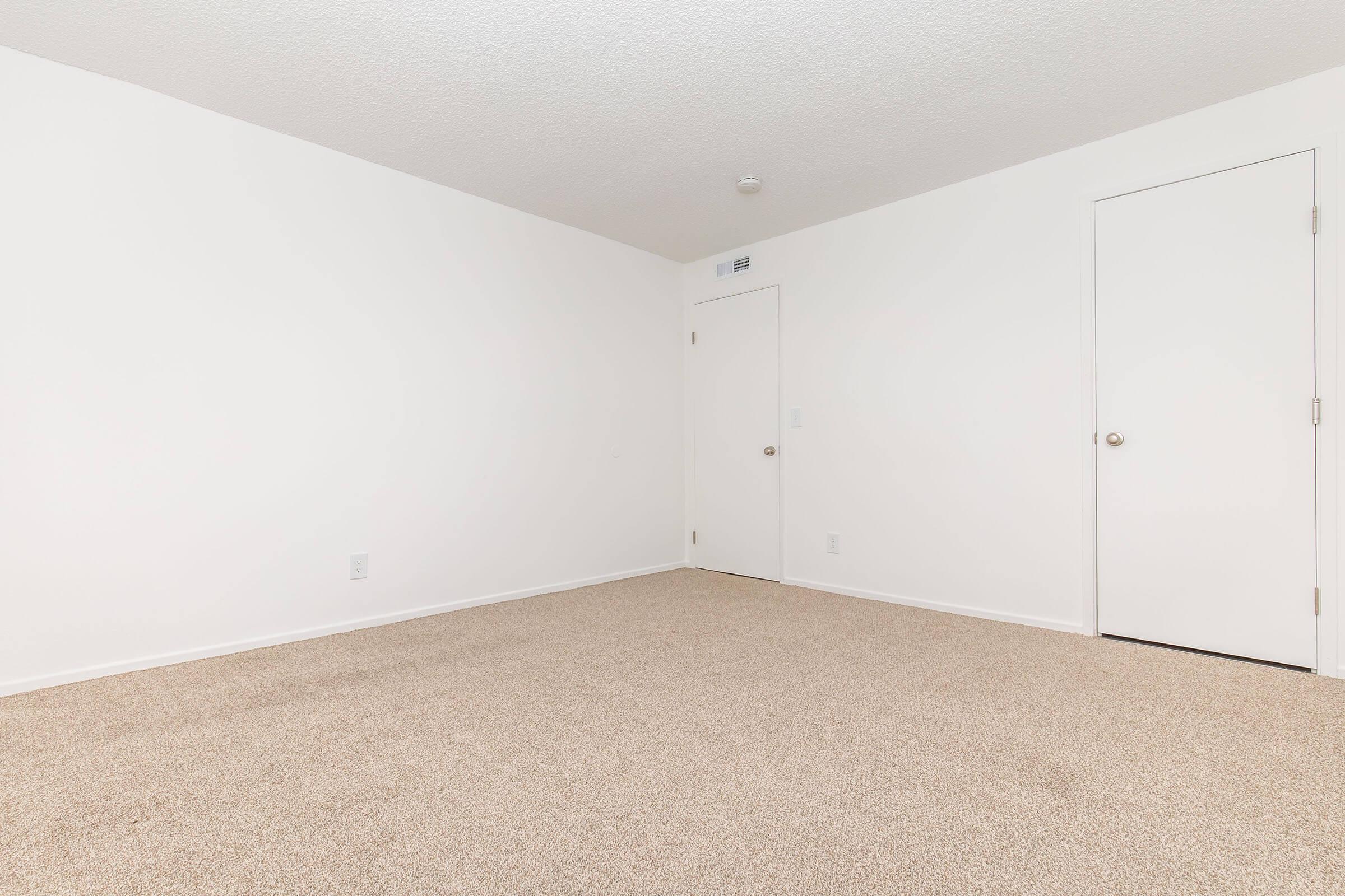 Empty room with light beige carpet and white walls. There is a closed door on the right and a wall with no furniture or decorations. The ceiling is plain with texture, and there is a small light fixture. Overall, the space is bright and minimalistic.