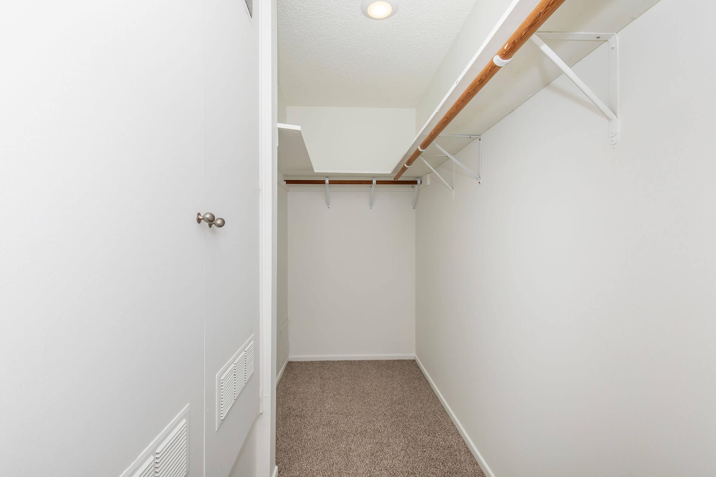 A small, empty closet featuring white walls and a carpeted floor. There is a wooden rod for hanging clothes and several metal shelves along the walls. Soft overhead lighting illuminates the space, creating a bright and clean appearance.