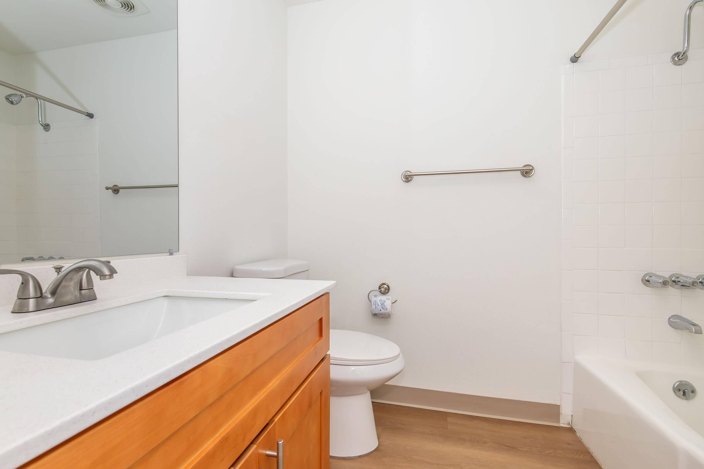 A bright, clean bathroom featuring a white sink with a modern faucet, a wooden cabinet beneath, a toilet, and a bathtub with a showerhead. The walls are light-colored, and there is a large mirror above the sink. A hand towel is hanging on a bar near the toilet, and the floor is finished with light wood.