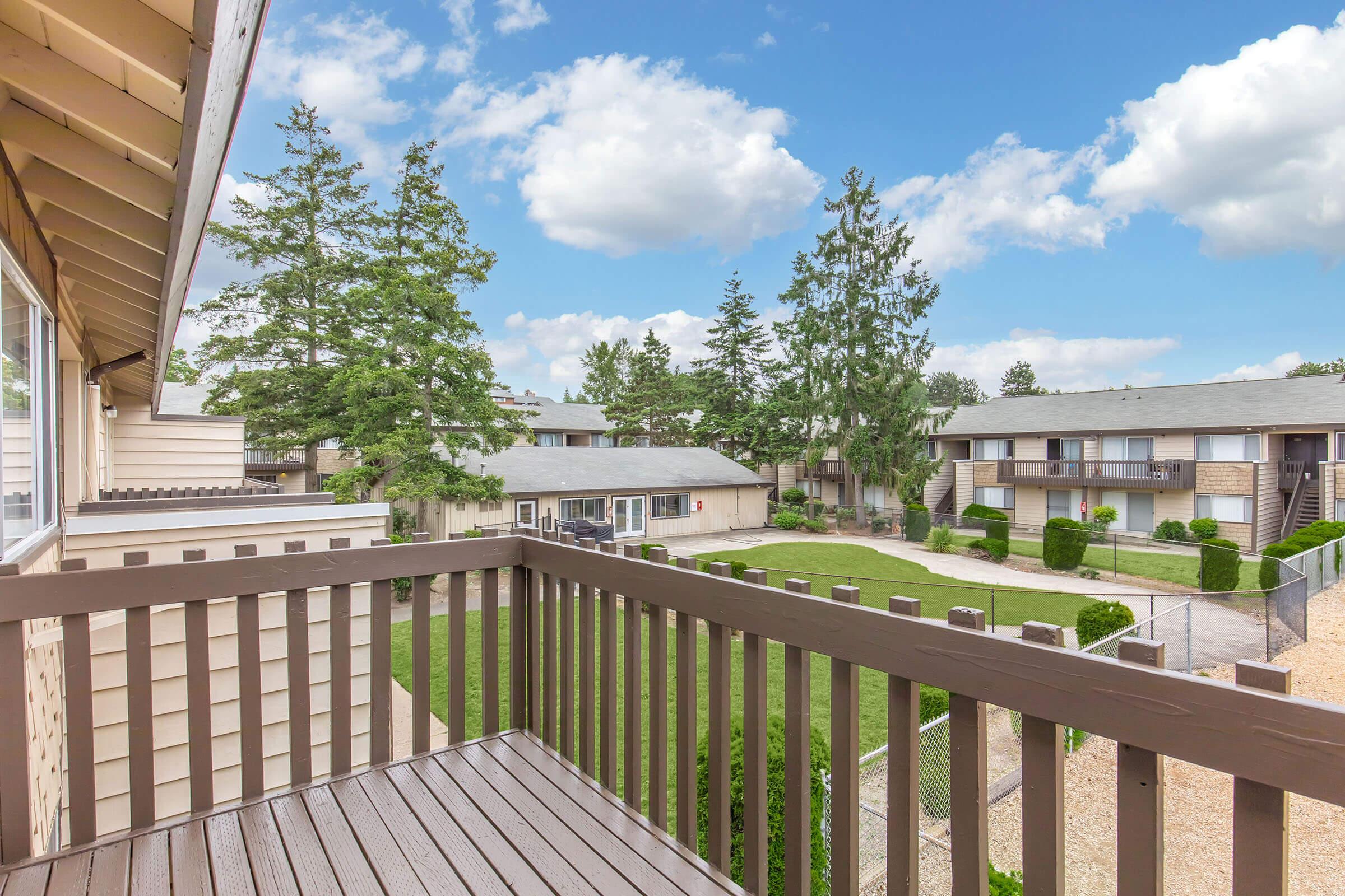 View from a balcony overlooking a landscaped courtyard with green grass and well-maintained shrubs. In the background, there are multiple buildings surrounded by trees under a partly cloudy sky.