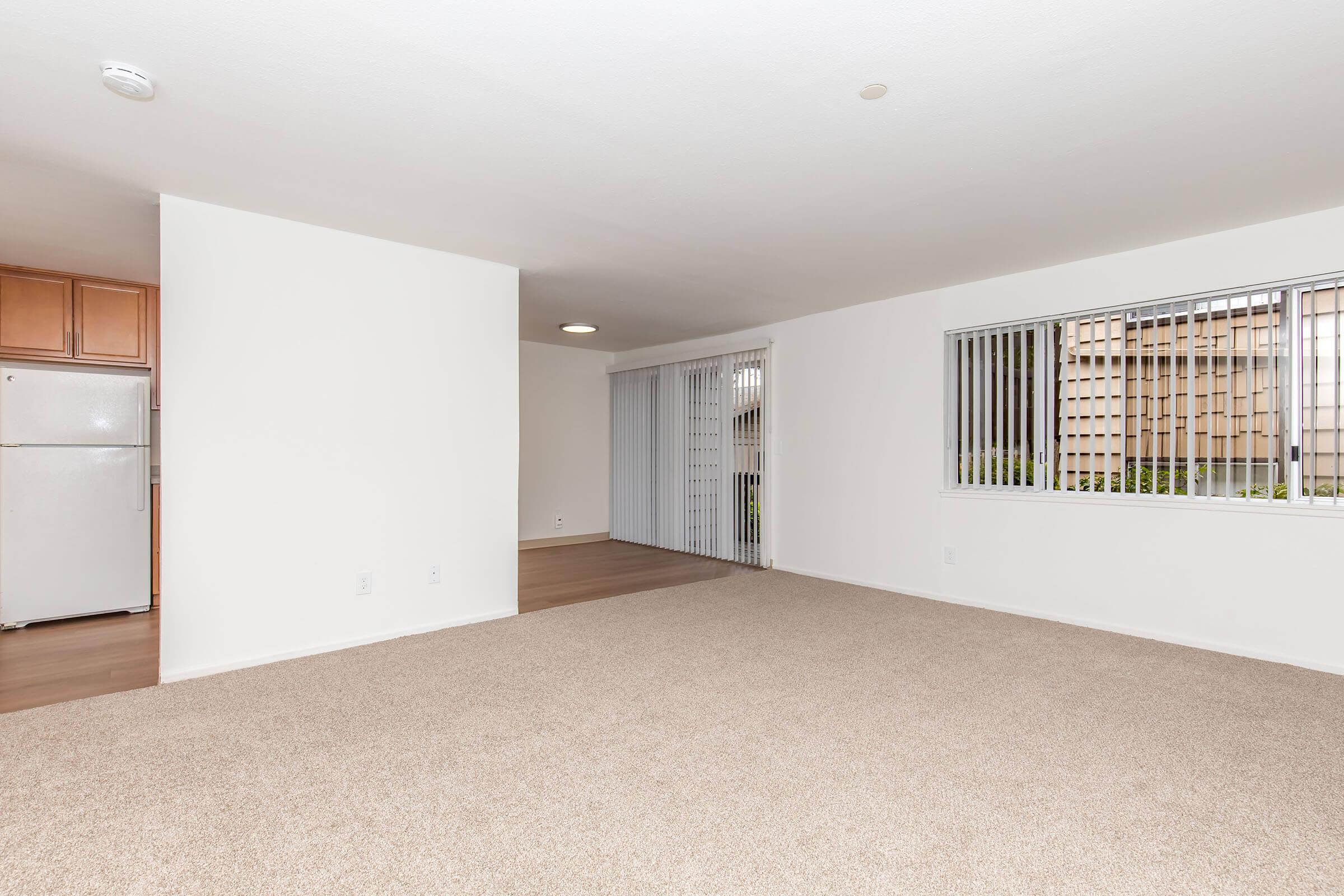 A spacious, empty living room with beige carpet, white walls, and large windows featuring vertical blinds. On the left, there is a kitchen area with a refrigerator visible. There is natural light coming in, creating a bright and airy atmosphere.