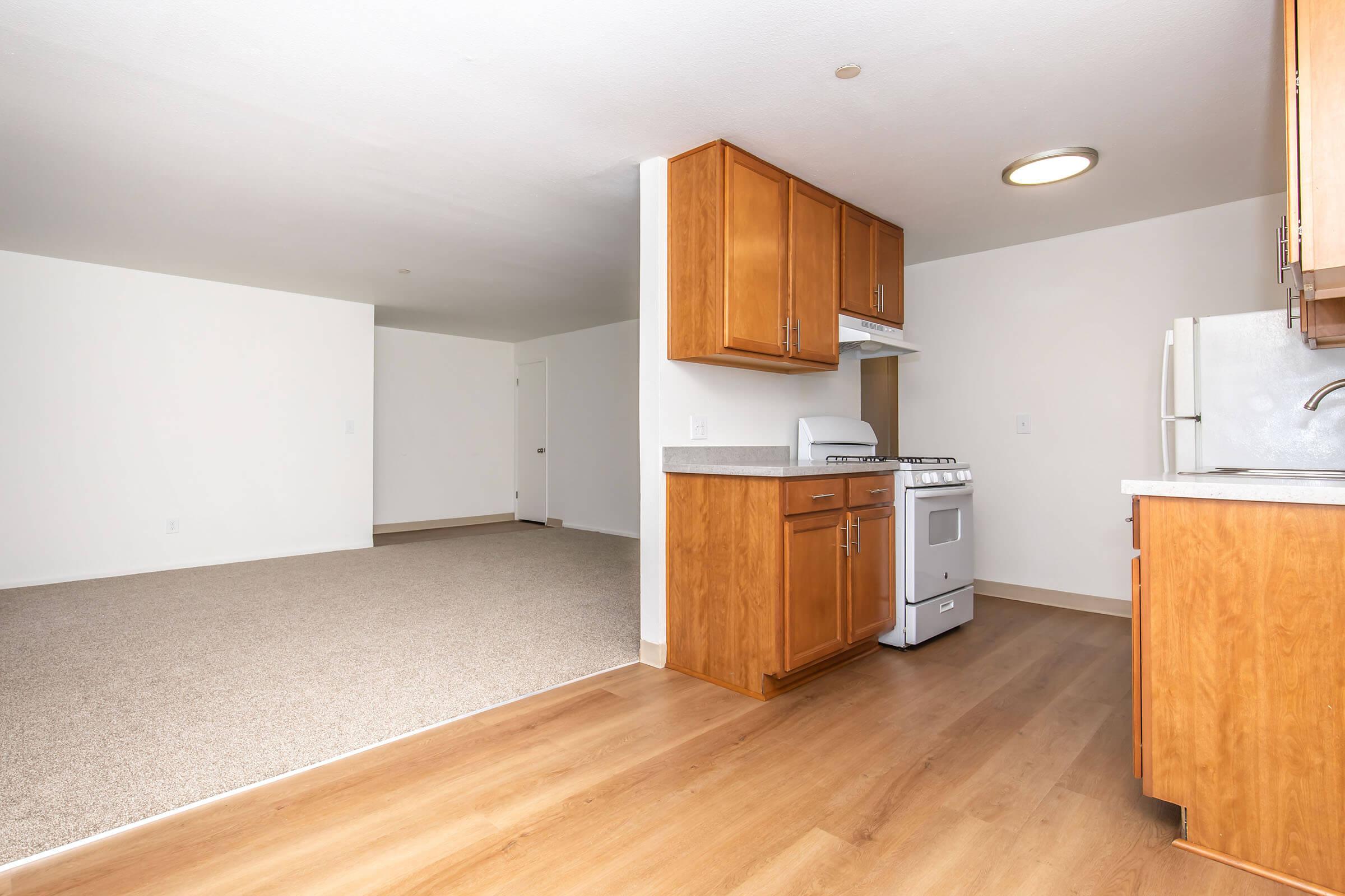 A modern kitchen featuring wooden cabinets, a white stove, and a countertop. The background shows an open living space with neutral-colored carpet and walls, illuminated by natural light. The layout suggests a spacious and inviting apartment.