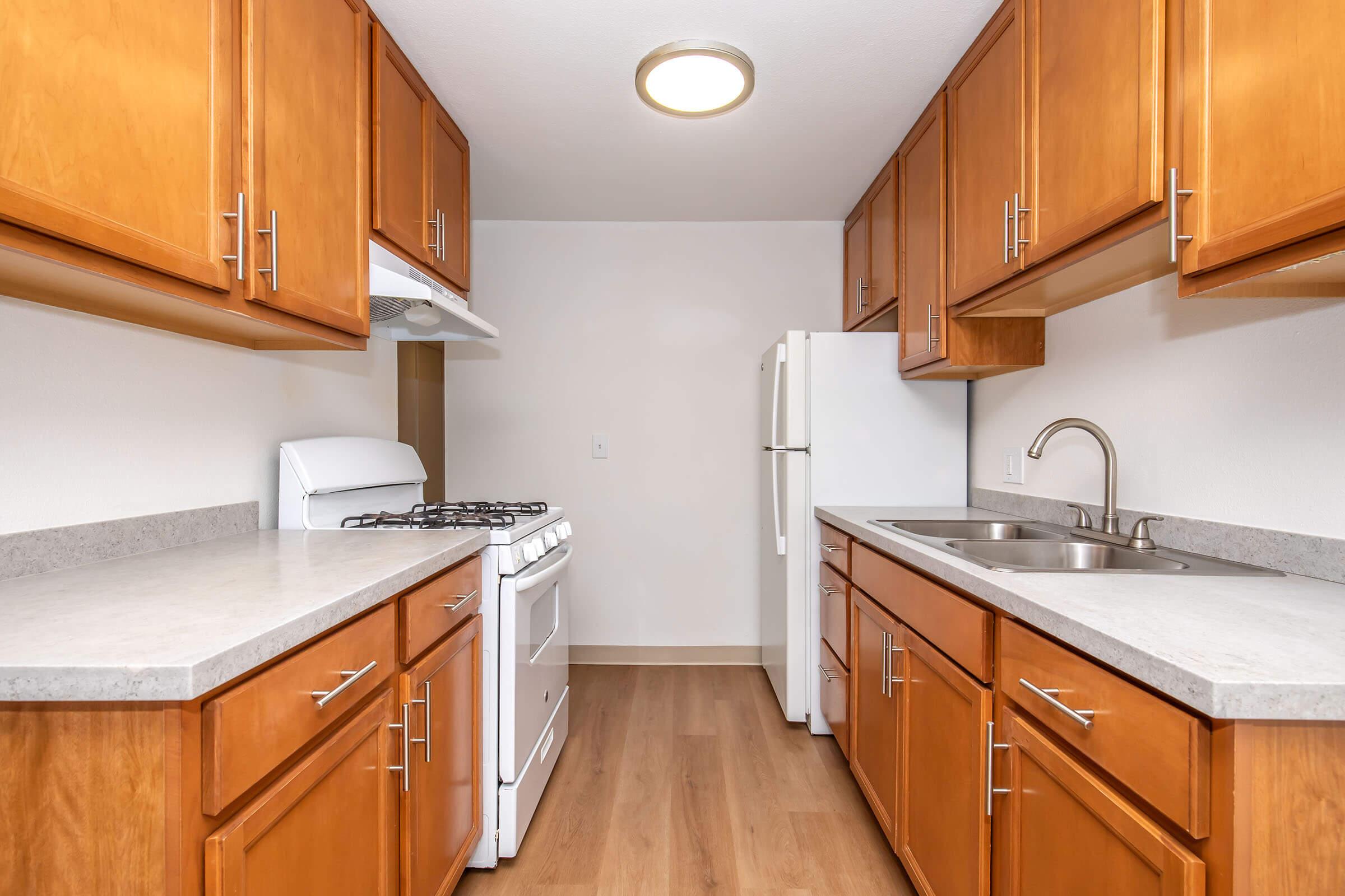 A modern kitchen featuring wooden cabinets, a gas stove, a white refrigerator, and a stainless steel sink. The countertops are light, and the flooring is wood-like. The walls are painted a neutral color, creating a clean and warm atmosphere in the space.