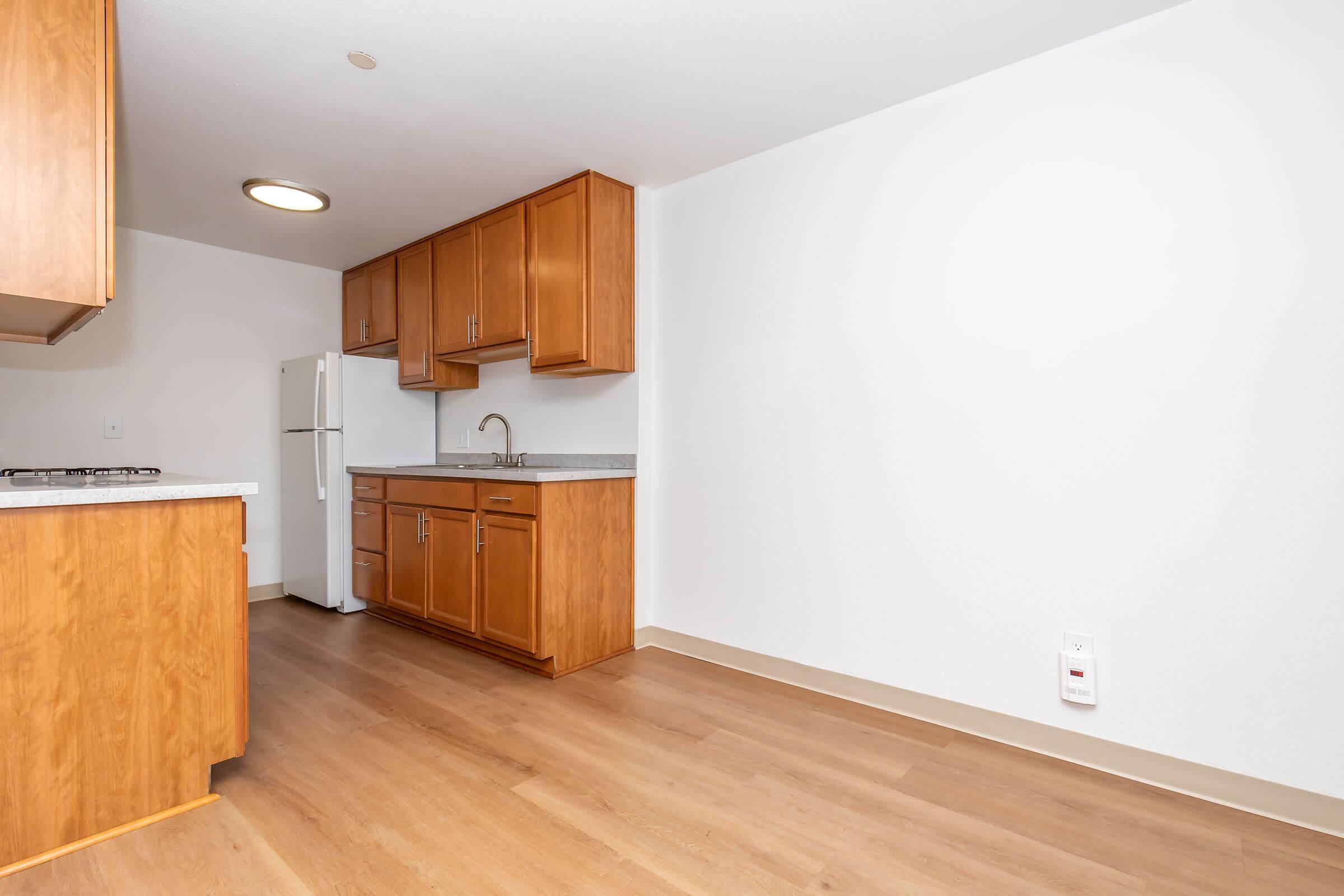 A modern kitchen featuring wooden cabinets, a white refrigerator, and a sink. The space has a hardwood floor and light-colored walls, with a large window allowing natural light. The kitchen area includes a countertop with a gas stove and minimal decor, creating a clean, functional look.