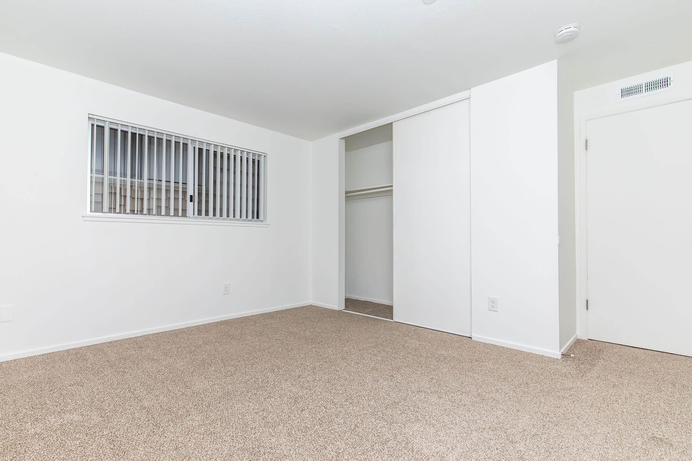 A spacious, empty room featuring light-colored walls and carpet. There's a window with blinds on one side and a closet with sliding doors on the other. The neutral decor allows for various design possibilities, making it suitable for different purposes such as a bedroom or office.
