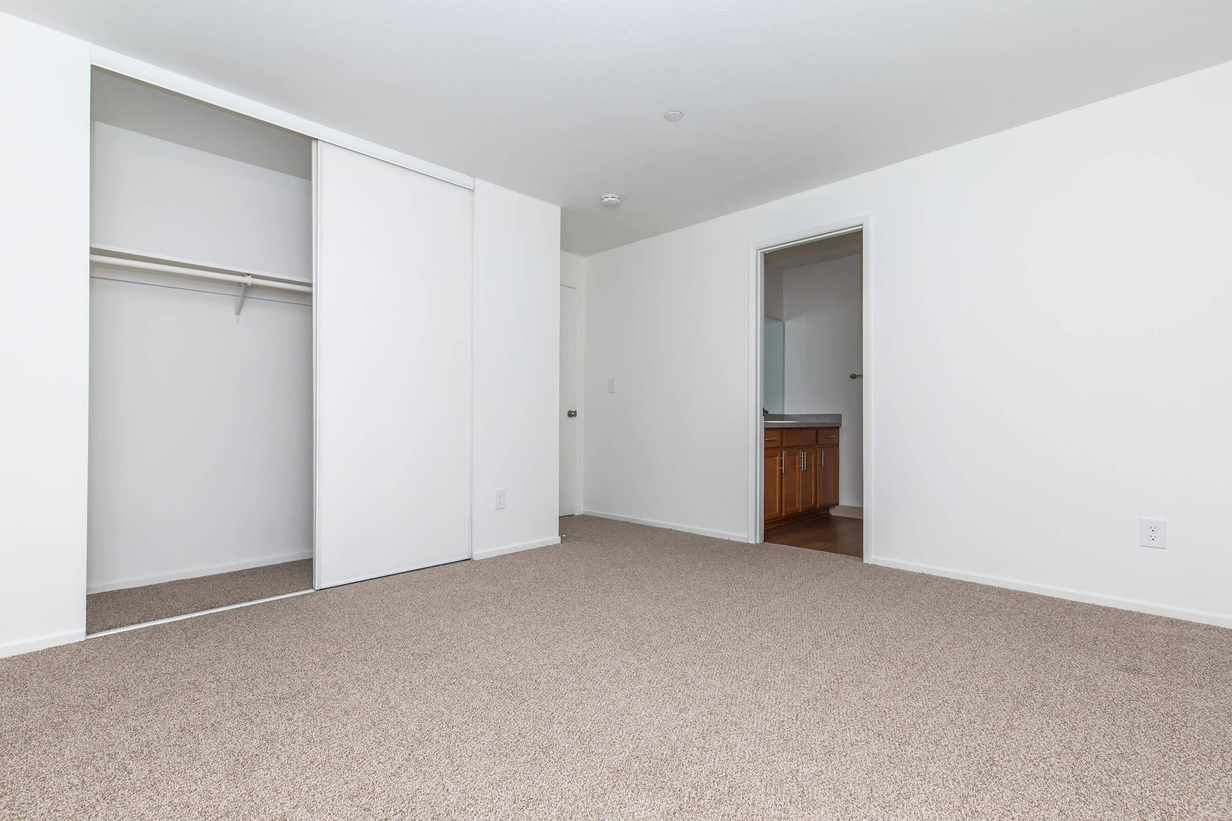 Empty room with light-colored walls and a tan carpet. There is a sliding closet with no doors open on the left side, and a doorway leading to another room on the right. The space is well-lit and has a clean, minimalistic aesthetic.