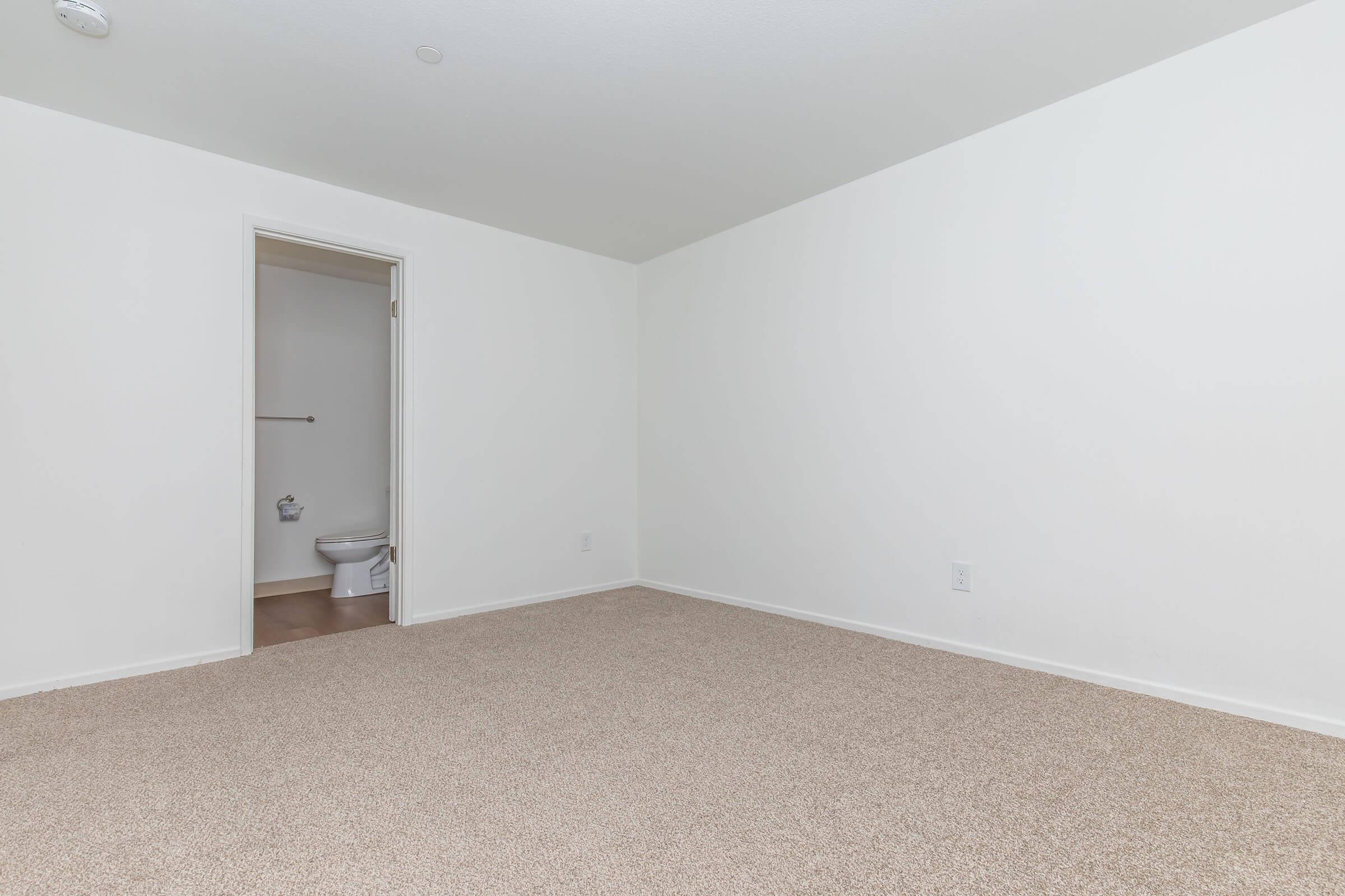 Empty room with beige carpeting and white walls. A closed door leads to a small bathroom area, visible from the side of the room. The space is well-lit and uncluttered, suggesting it could be used for various purposes. There are no furnishings or decorations present.