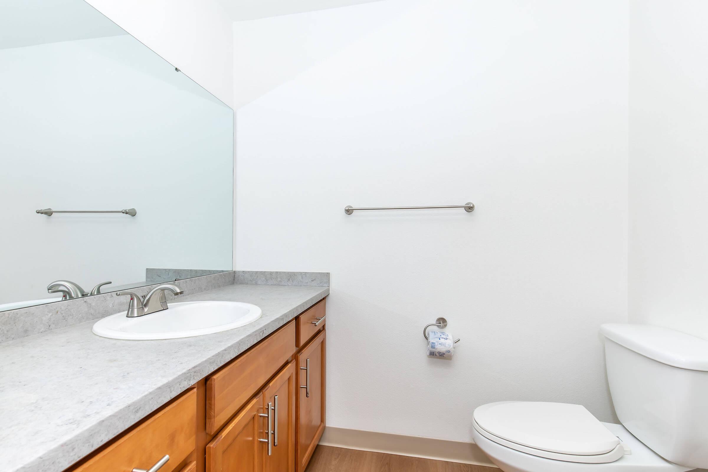 A clean, modern bathroom featuring a wide mirror above a gray countertop with a single sink, wooden cabinetry, a toilet on the right, and a towel bar on the wall. The walls are painted white and the flooring is light-colored, creating a bright and spacious atmosphere.