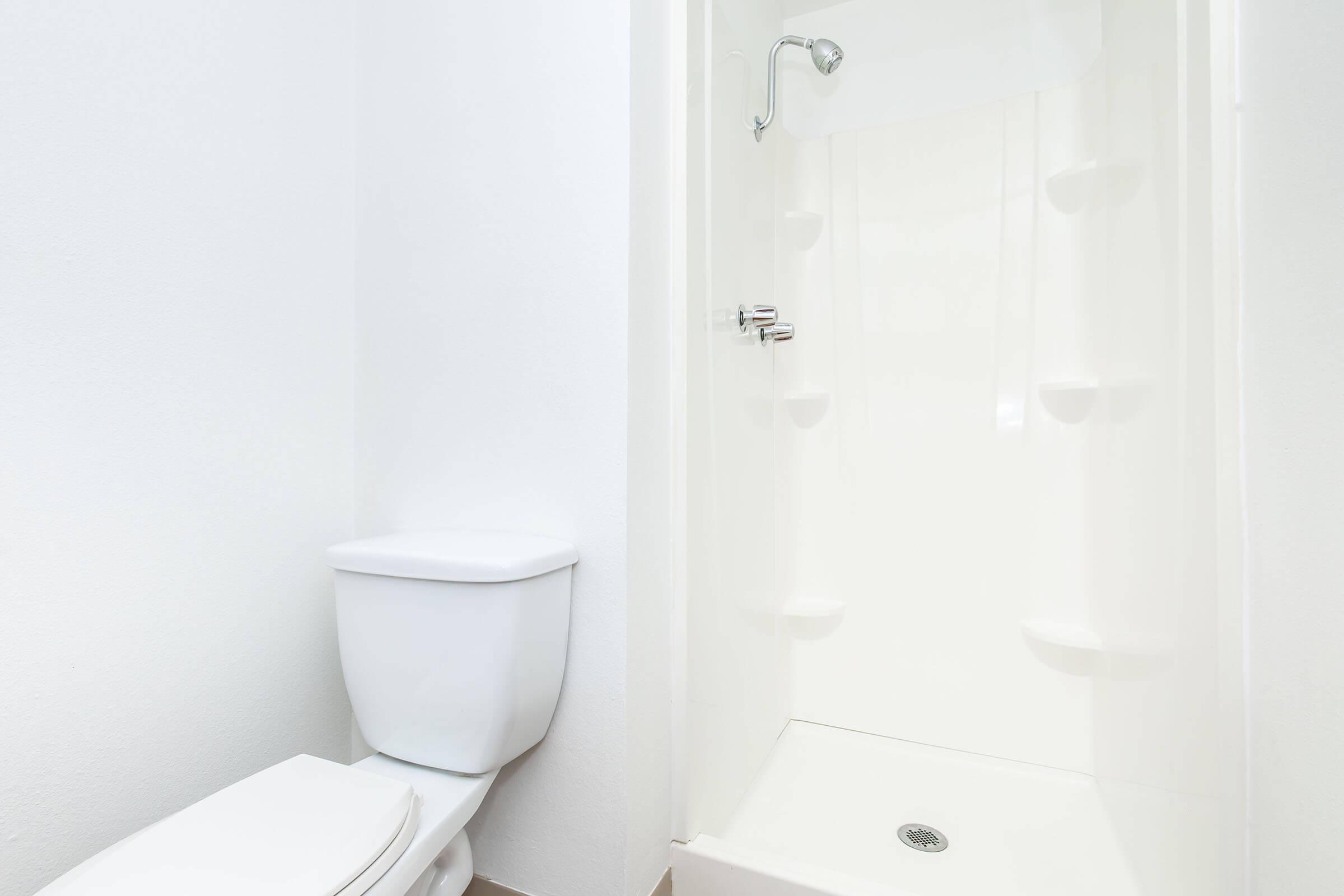 A clean bathroom featuring a white toilet and a small shower stall. The walls are painted white, enhancing the bright and minimalist aesthetic. The floor is simple, complementing the overall modern and tidy appearance of the space.