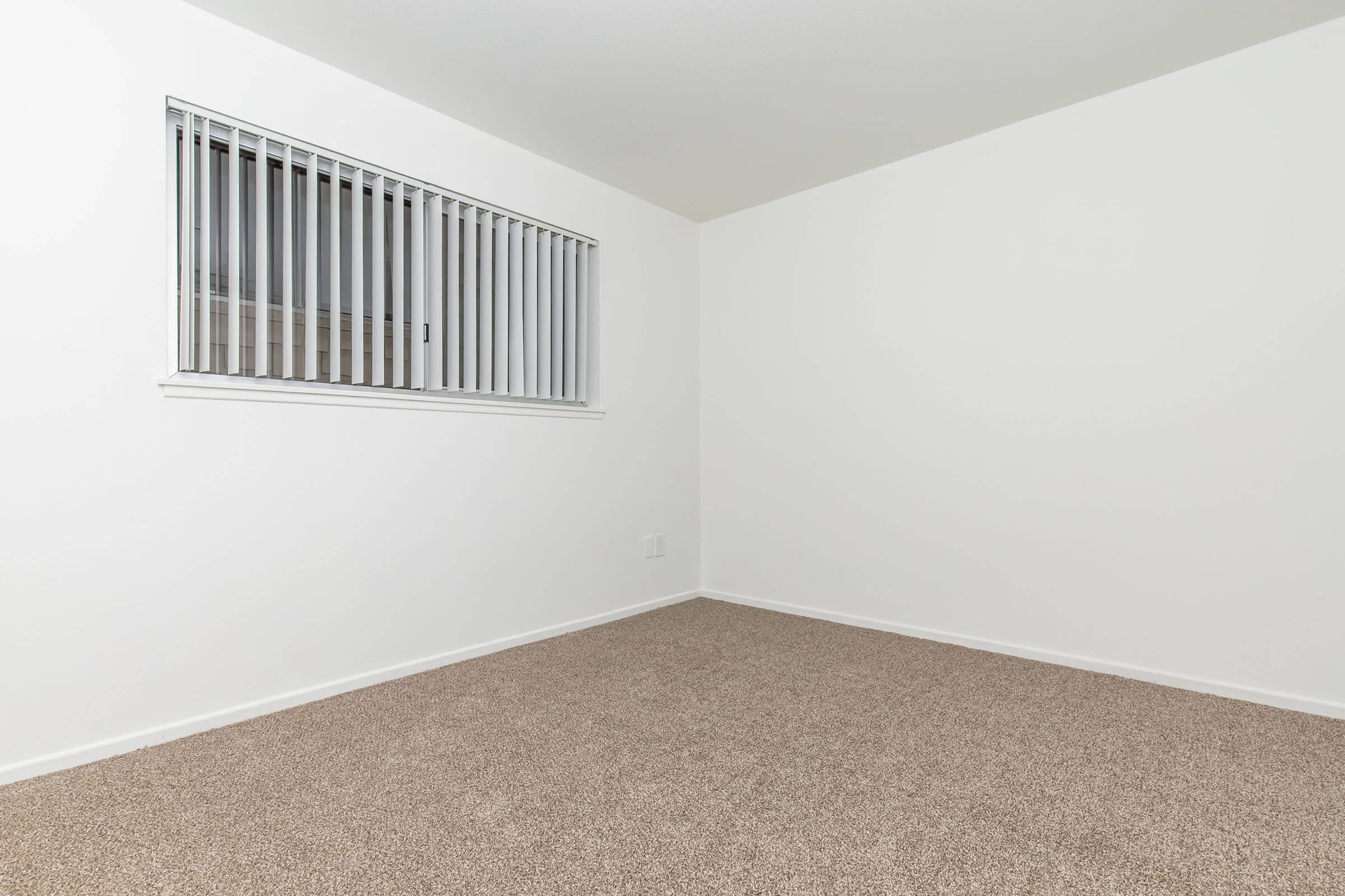 A minimalist room featuring light-colored walls and a carpeted floor. There's a window on the left side with vertical blinds, allowing natural light to enter. The space is largely empty, conveying a sense of simplicity and openness.