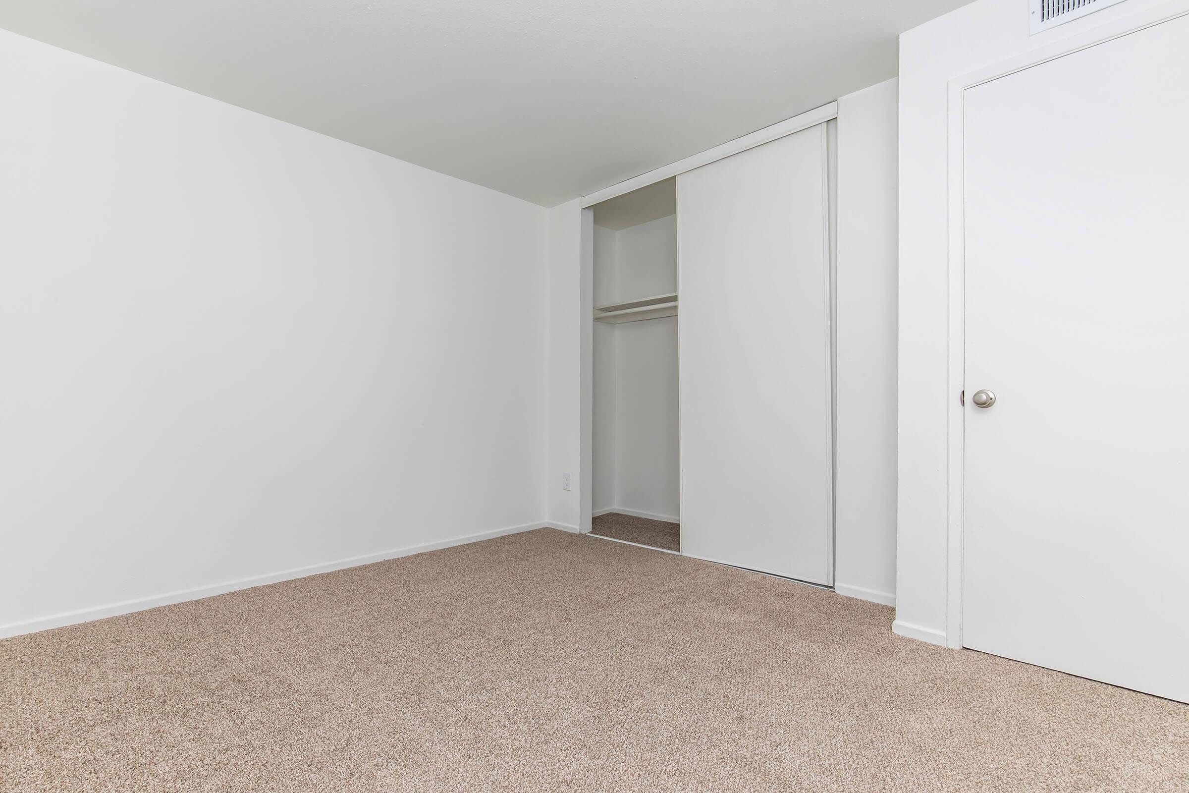 A sparsely furnished room with beige carpet and white walls. There's a sliding closet door on the left side and a closed door on the right. The room appears empty and well-lit, creating a spacious feel.