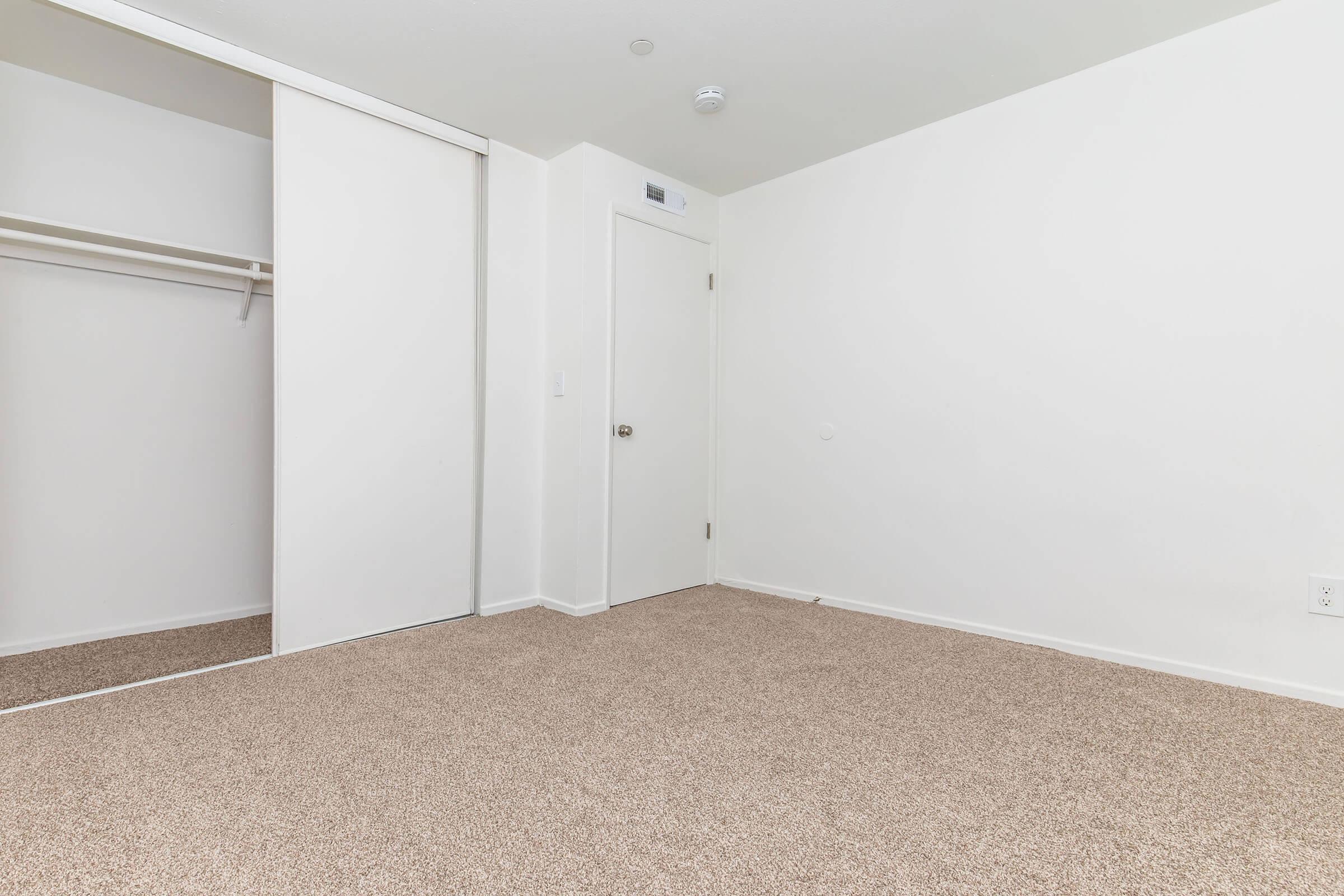 A minimalist, empty room featuring light beige carpet, white walls, and a closet with sliding doors. A plain door is visible on the right side, and there are no furnishings or decorations in the space, creating a clean and open feel.