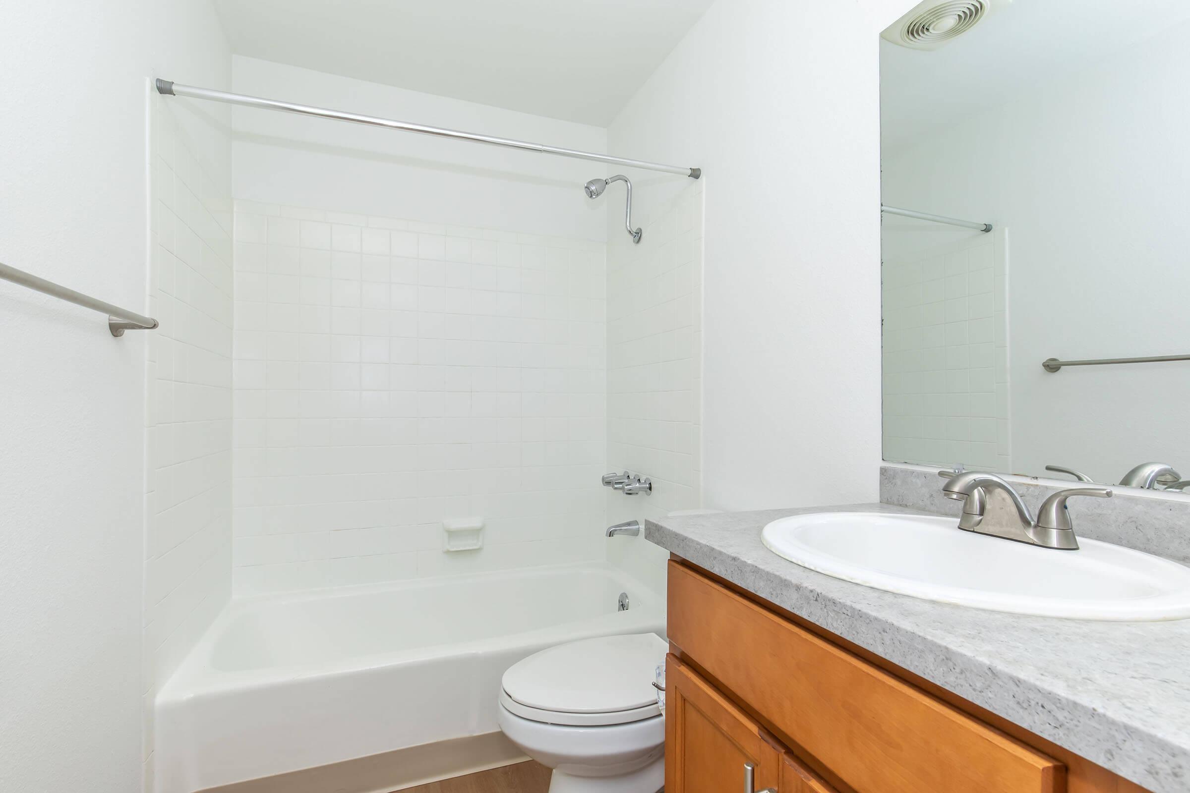 A clean and modern bathroom featuring a white tiled shower/bathtub combination, a single sink with a granite countertop, and wooden cabinet storage. The walls are painted white, and the floor is light-colored wood, creating a bright and spacious atmosphere.
