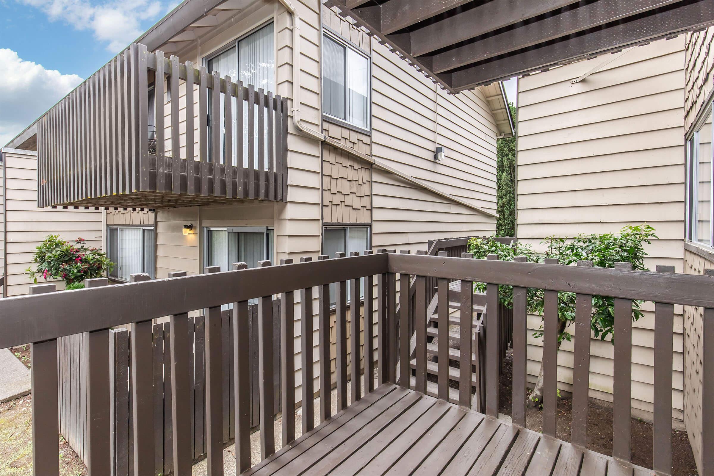 View of a wooden balcony surrounded by beige apartment buildings. The balcony features wooden railings and overlooks a stairway. Green plants are visible near the base of the building, and there are large windows on the upper floors. The sky is partly cloudy in the background.