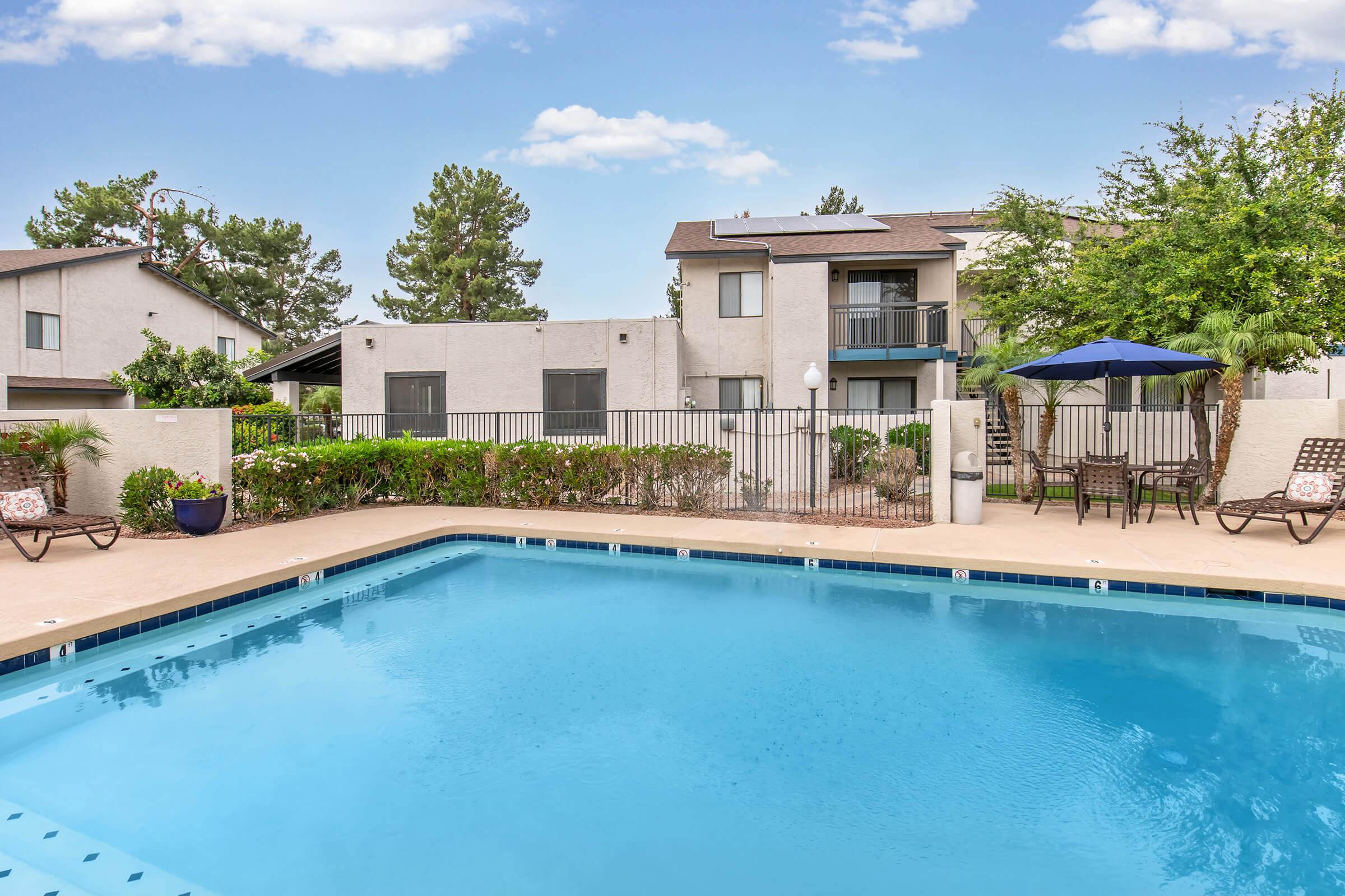 A clean, inviting swimming pool surrounded by lounge chairs and a small table with an umbrella. In the background, there are two multi-story residential buildings with balconies and landscaped greenery, under a clear blue sky.