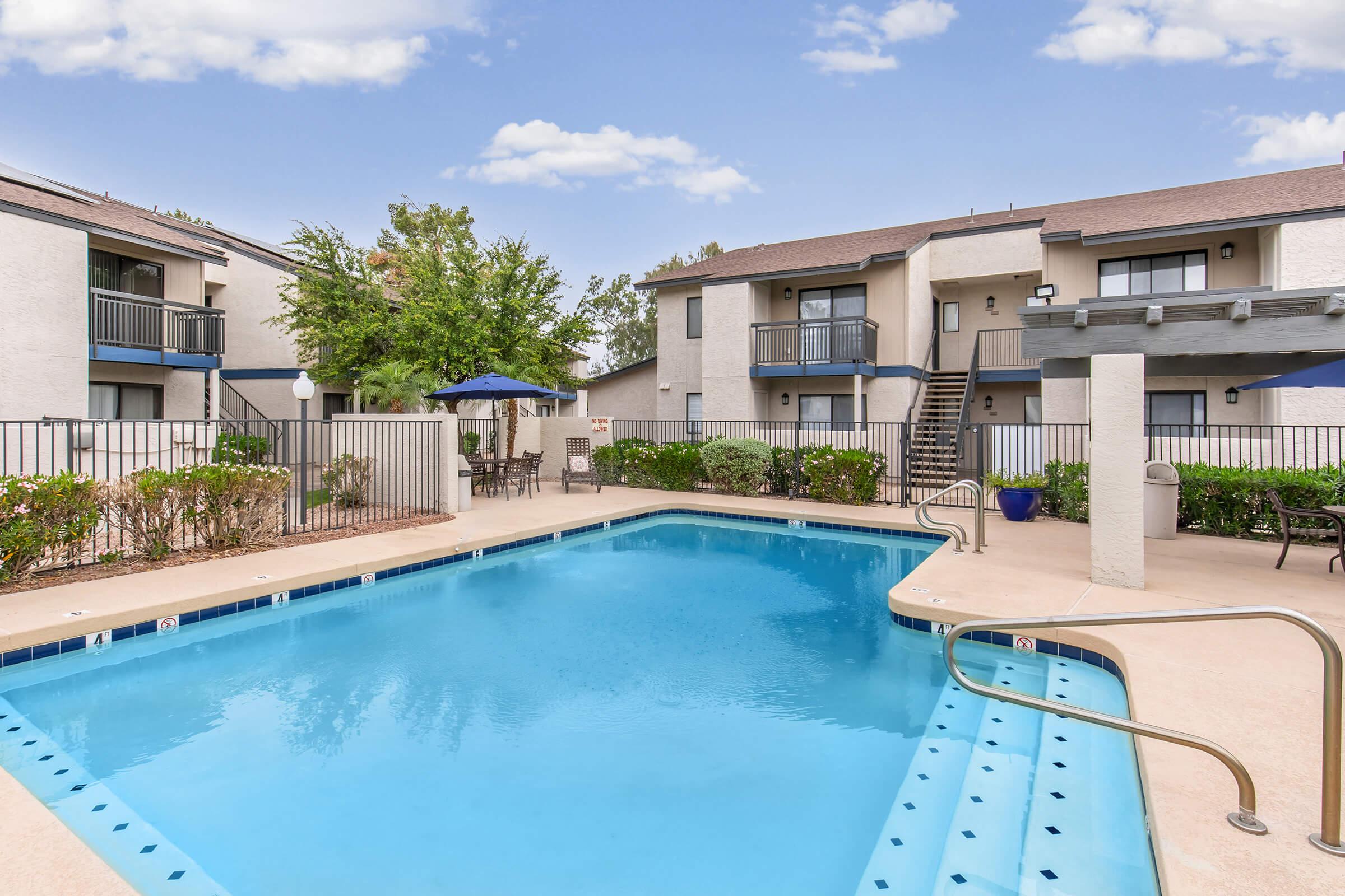 A well-maintained outdoor swimming pool surrounded by a patio area and landscaped greenery. Two nearby apartment buildings are visible, with balconies overlooking the pool. Blue skies and a few clouds create a bright, inviting atmosphere.