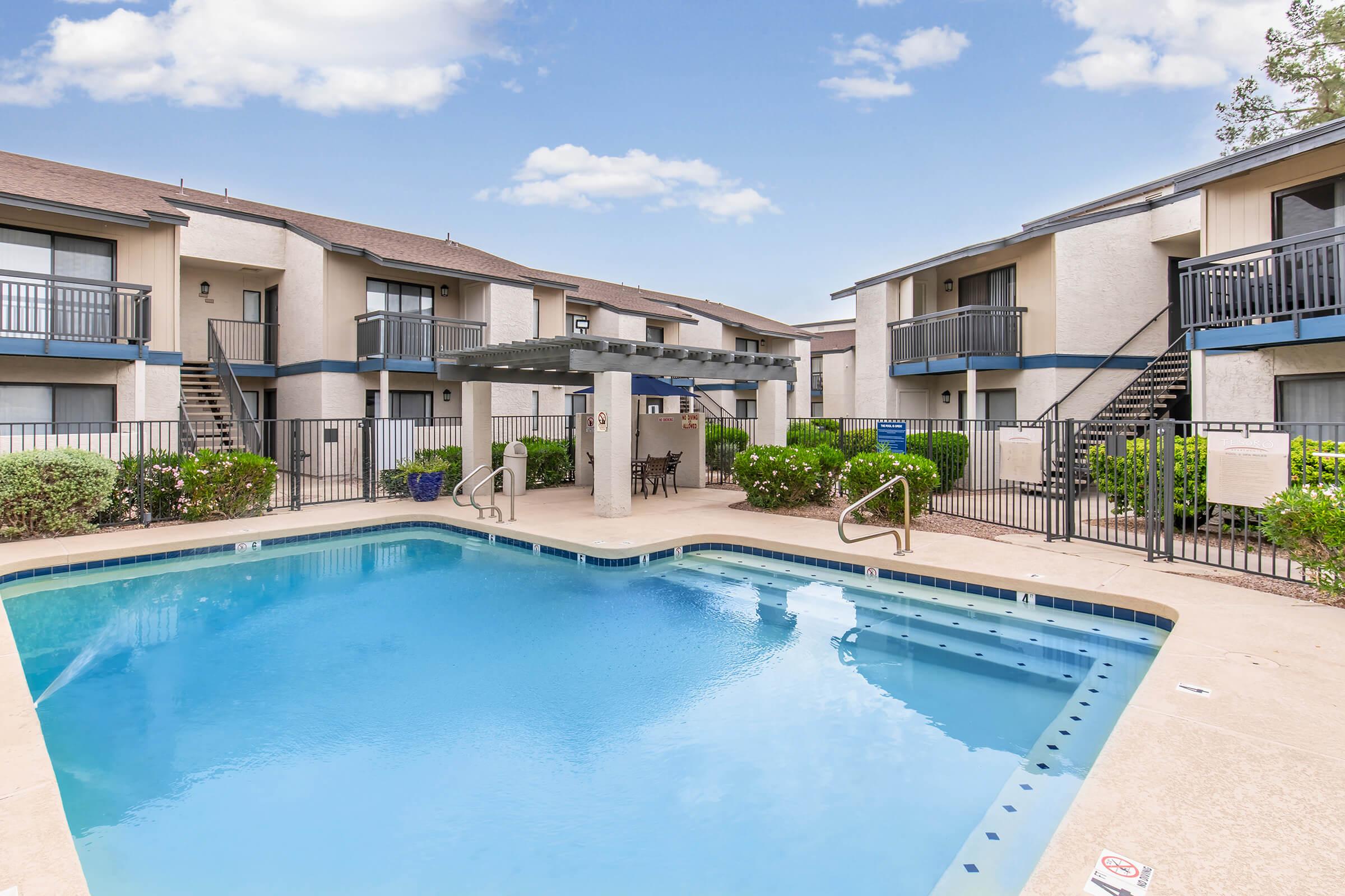 A tranquil pool area surrounded by landscaped greenery and two-story apartment buildings. The pool features a jacuzzi and lounge chairs, with a shaded sitting area nearby. Clear blue skies and fluffy clouds create a bright, inviting atmosphere.
