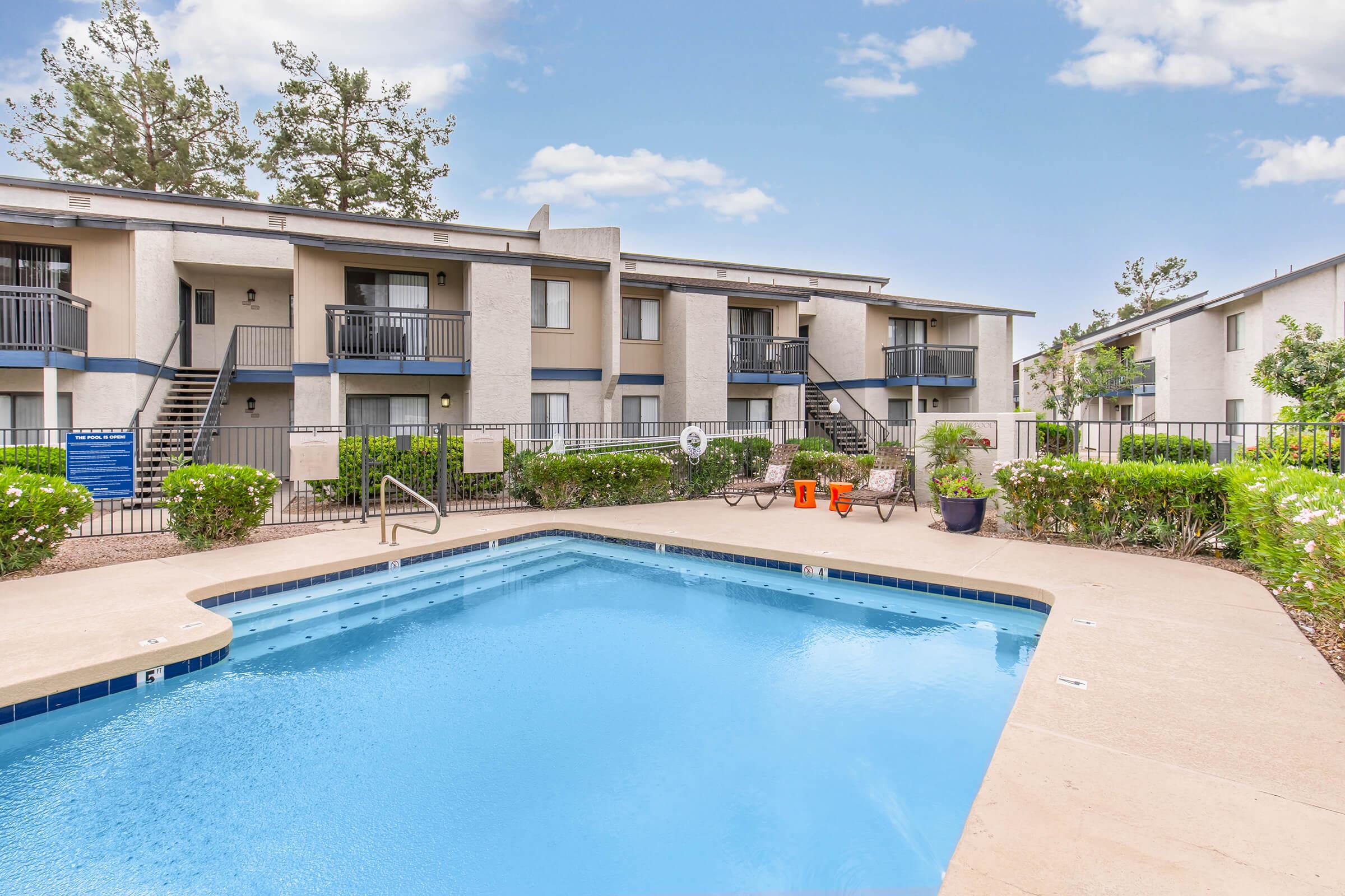 A view of an apartment complex with a swimming pool in the foreground. Two lounge chairs are placed near the pool, surrounded by well-maintained greenery and flowers. The buildings are two stories tall with balconies, and there are stairs leading to the upper level. The sky is clear and blue.