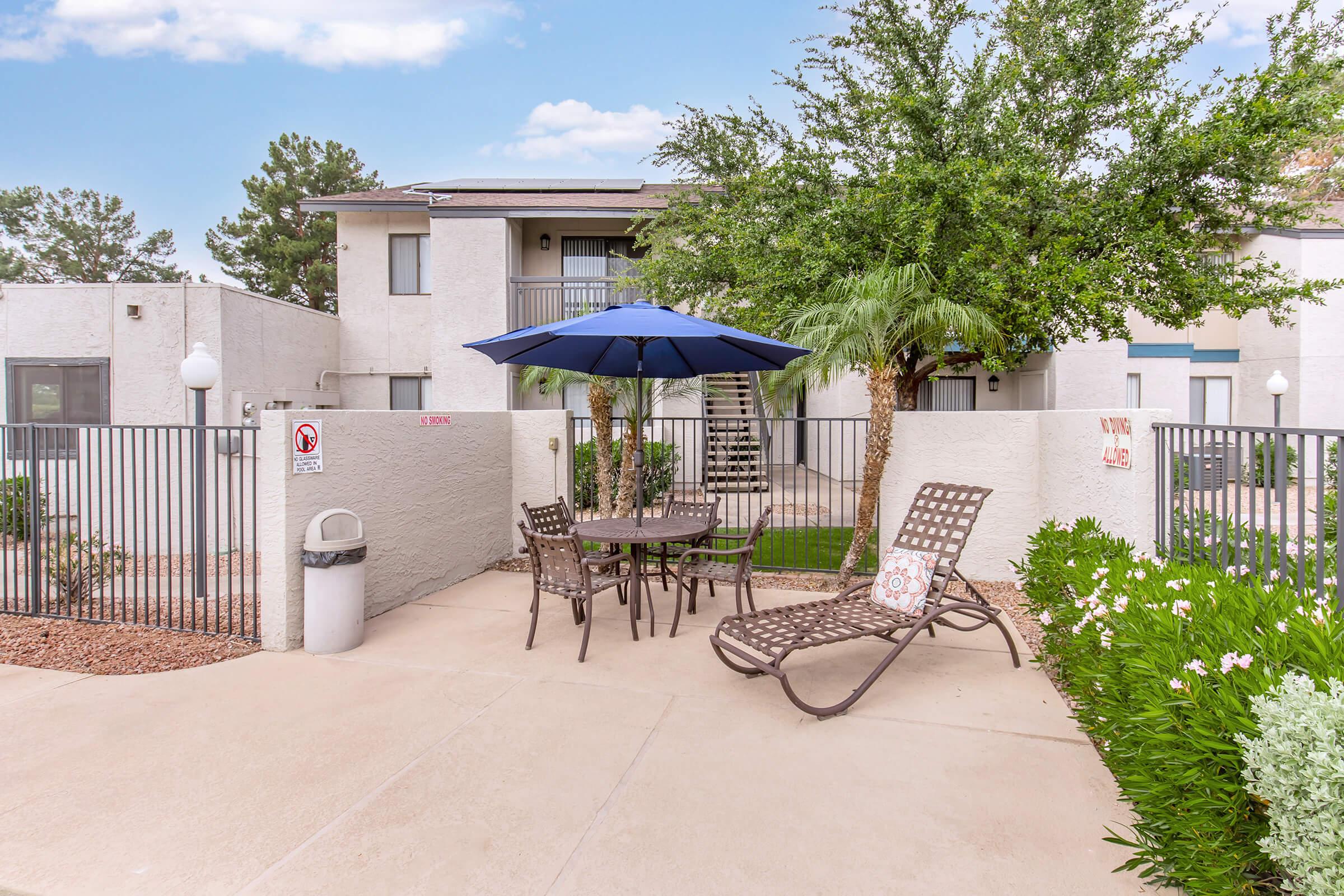 A tranquil outdoor seating area featuring a table with four chairs under a blue umbrella, surrounded by greenery and flower beds. A trash bin is nearby, and a gated entrance is visible in the background, with a building and trees providing a pleasant backdrop.