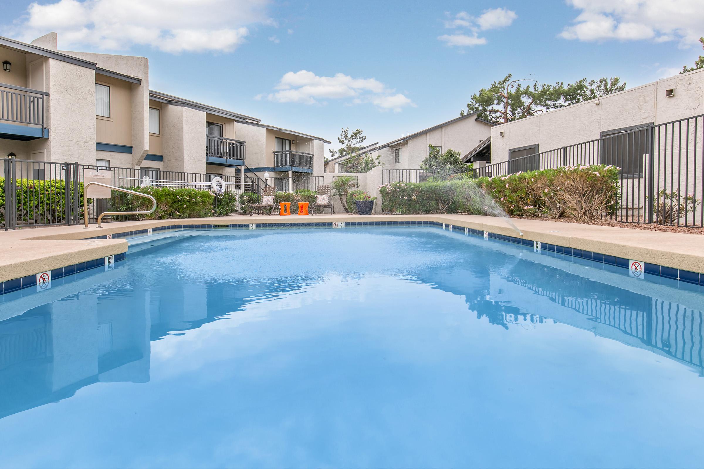 A clear, blue swimming pool surrounded by well-maintained landscaping and two-story apartment buildings in the background. The scene is bright and inviting, with a few clouds in the sky and lounge chairs near the pool area.
