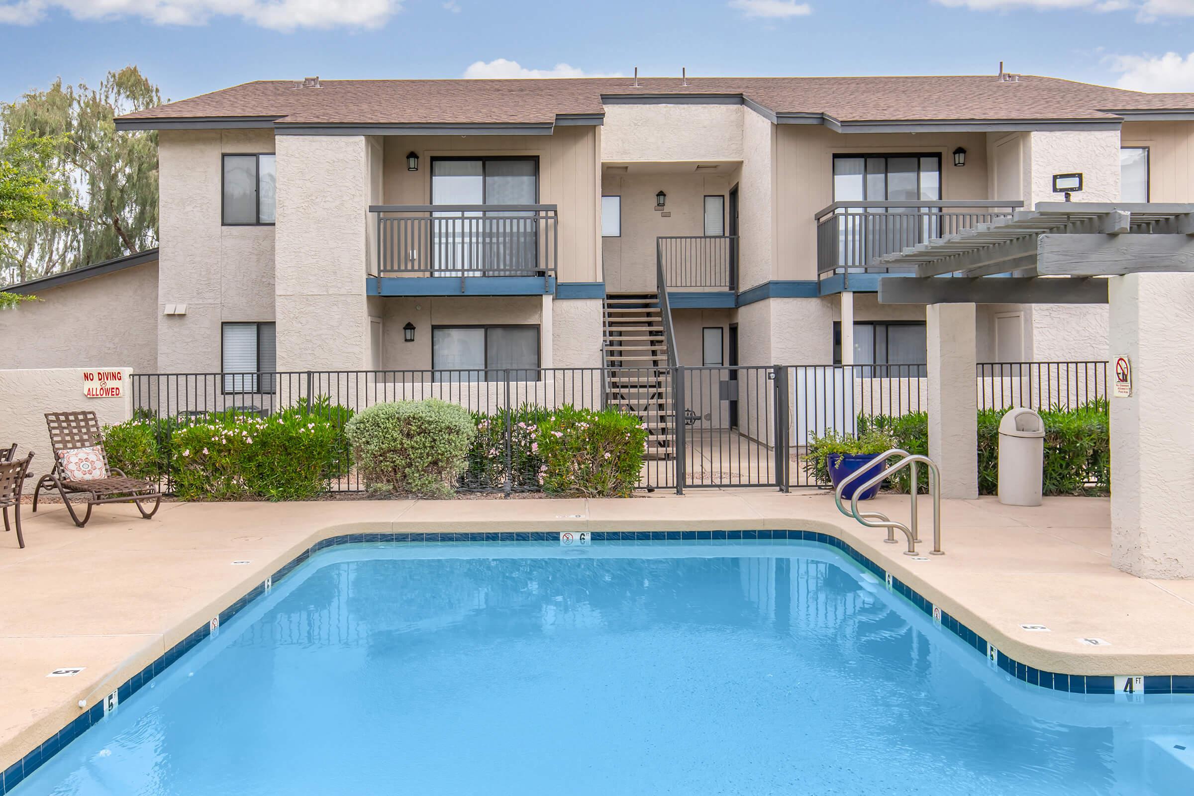 A view of an apartment complex featuring a two-story building with balconies overlooking a swimming pool. The pool area is surrounded by a fence and landscaped with shrubs. There is a staircase leading to the upper floor, and a sign indicating no diving. The sky is clear with a few clouds.
