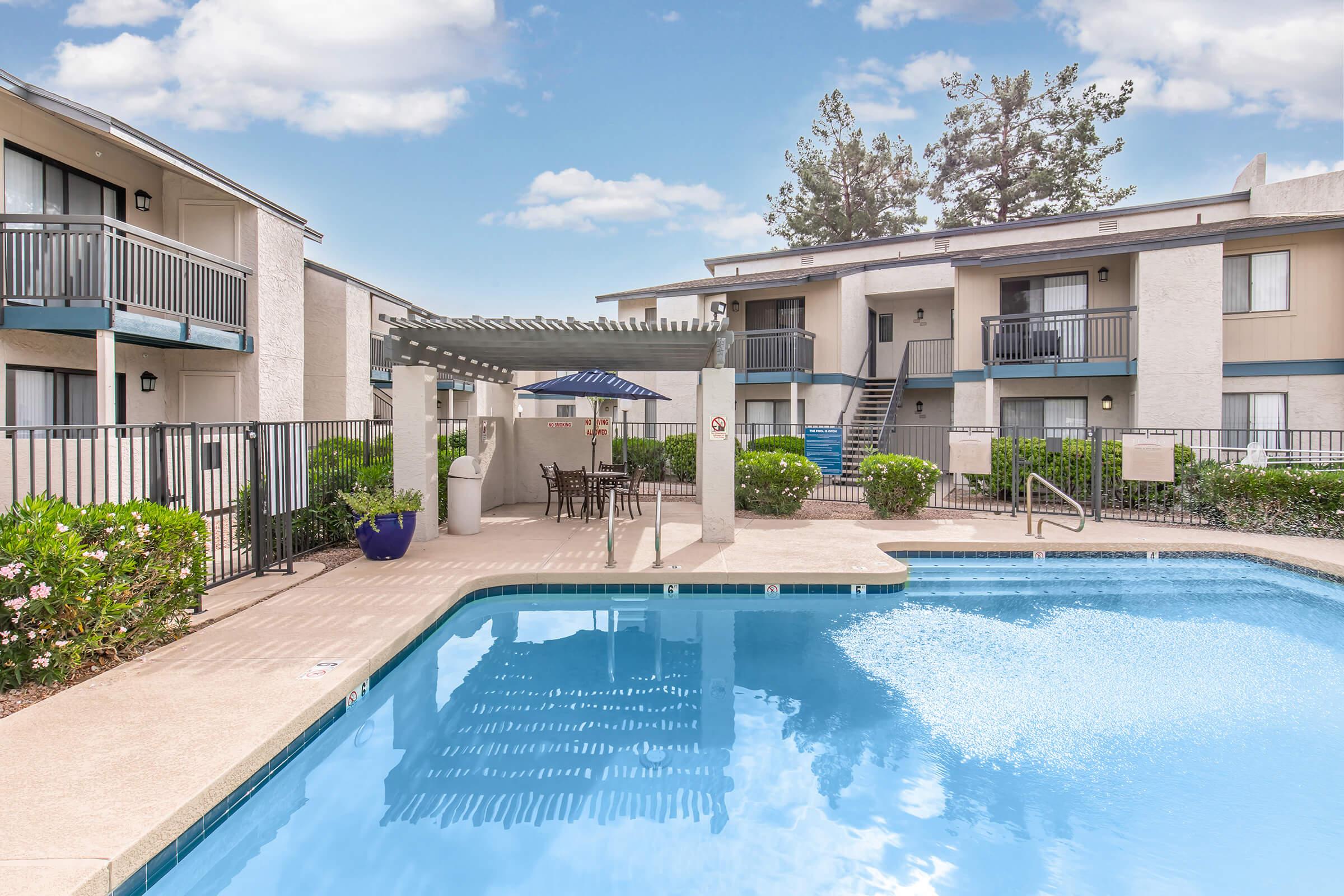 A clear blue swimming pool surrounded by landscaped greenery and a seating area with tables and umbrellas. In the background, there are two-story apartment buildings with balconies. The sky is bright with fluffy clouds.