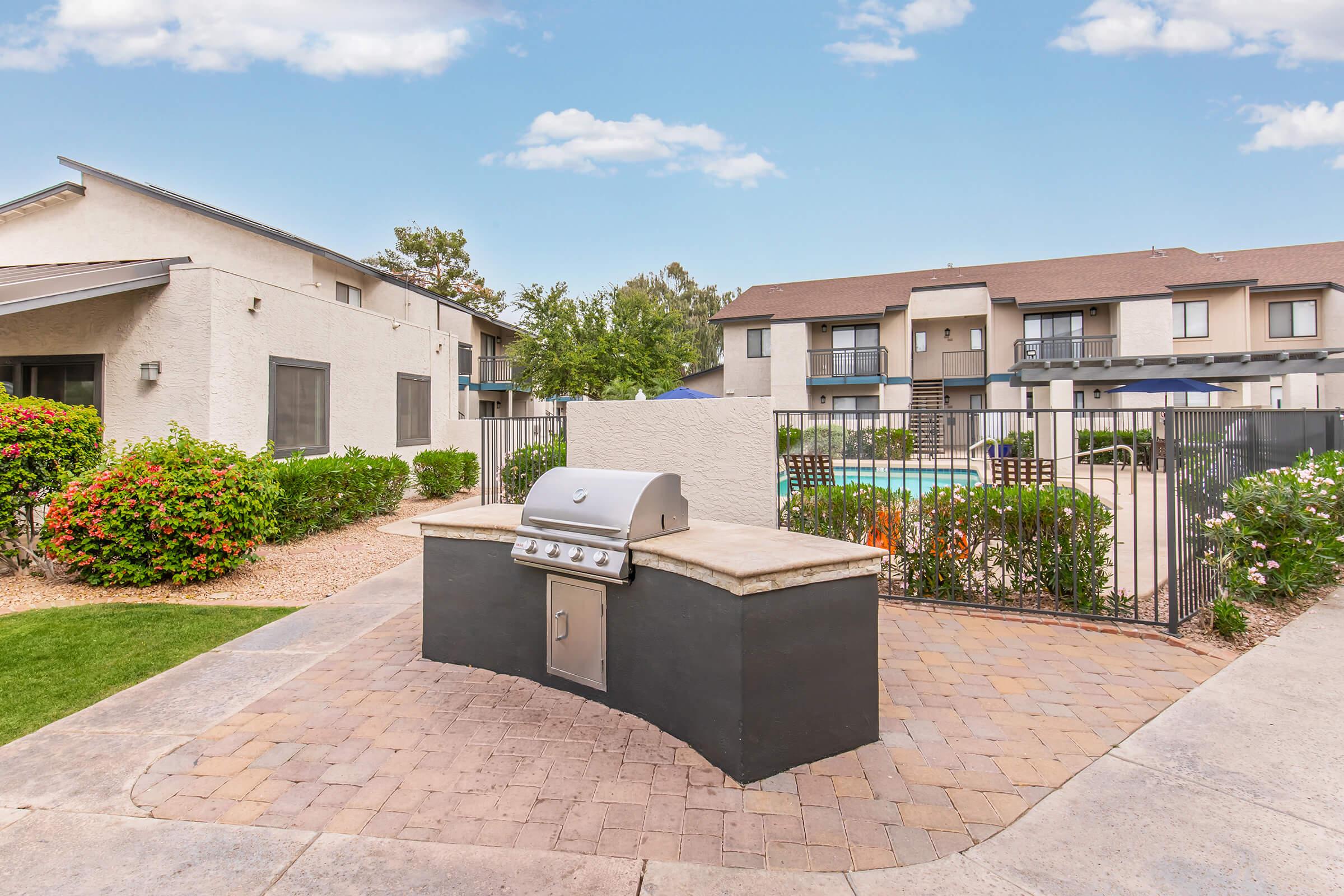 An outdoor barbecue grill setup on a stone patio, surrounded by lush green shrubs and flowers. In the background, there is a swimming pool area with several lounge chairs, bordered by a fence, and apartment buildings visible. The sky is clear and blue, creating a welcoming atmosphere.