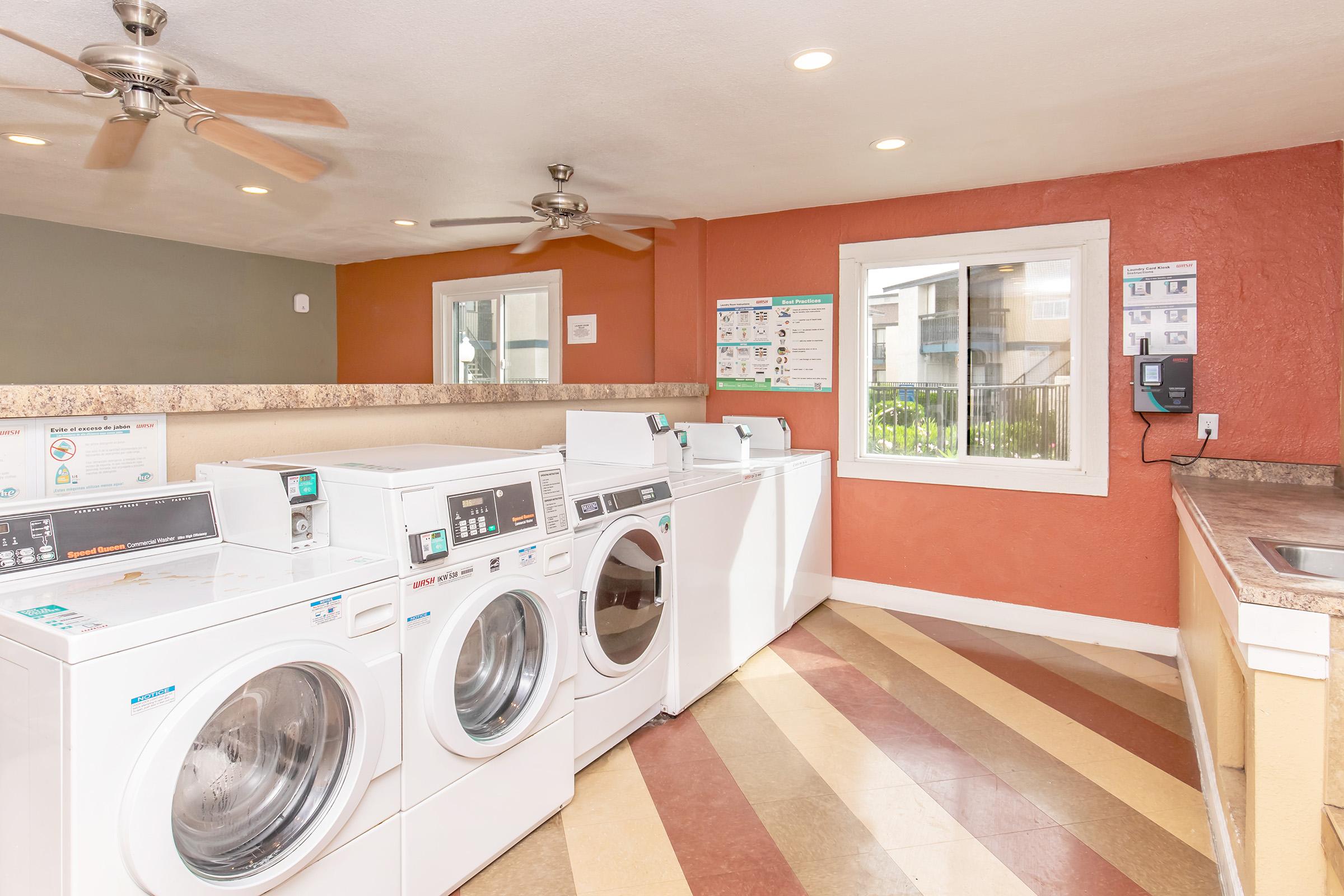 A clean and modern laundry room featuring several white washing machines and dryers. There are ceiling fans for ventilation, a large window allowing natural light, a countertop along one wall, and informational posters about laundry use displayed. The floor has a colorful, patterned design.