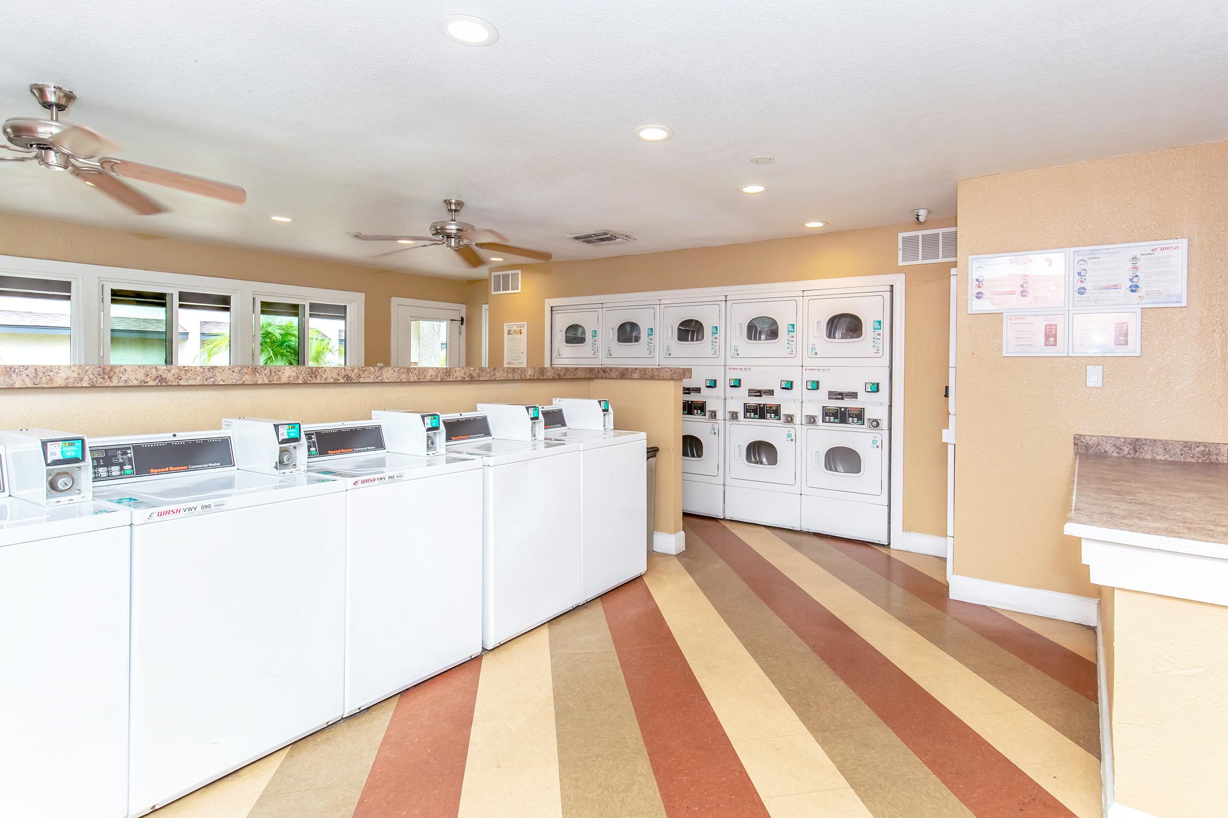 A well-lit laundry room featuring several white washing machines and dryers arranged neatly against the wall. The floor has a colorful, striped design. Windows provide natural light, and there are ceiling fans for ventilation. Nearby, instructions and safety information are displayed on the wall.