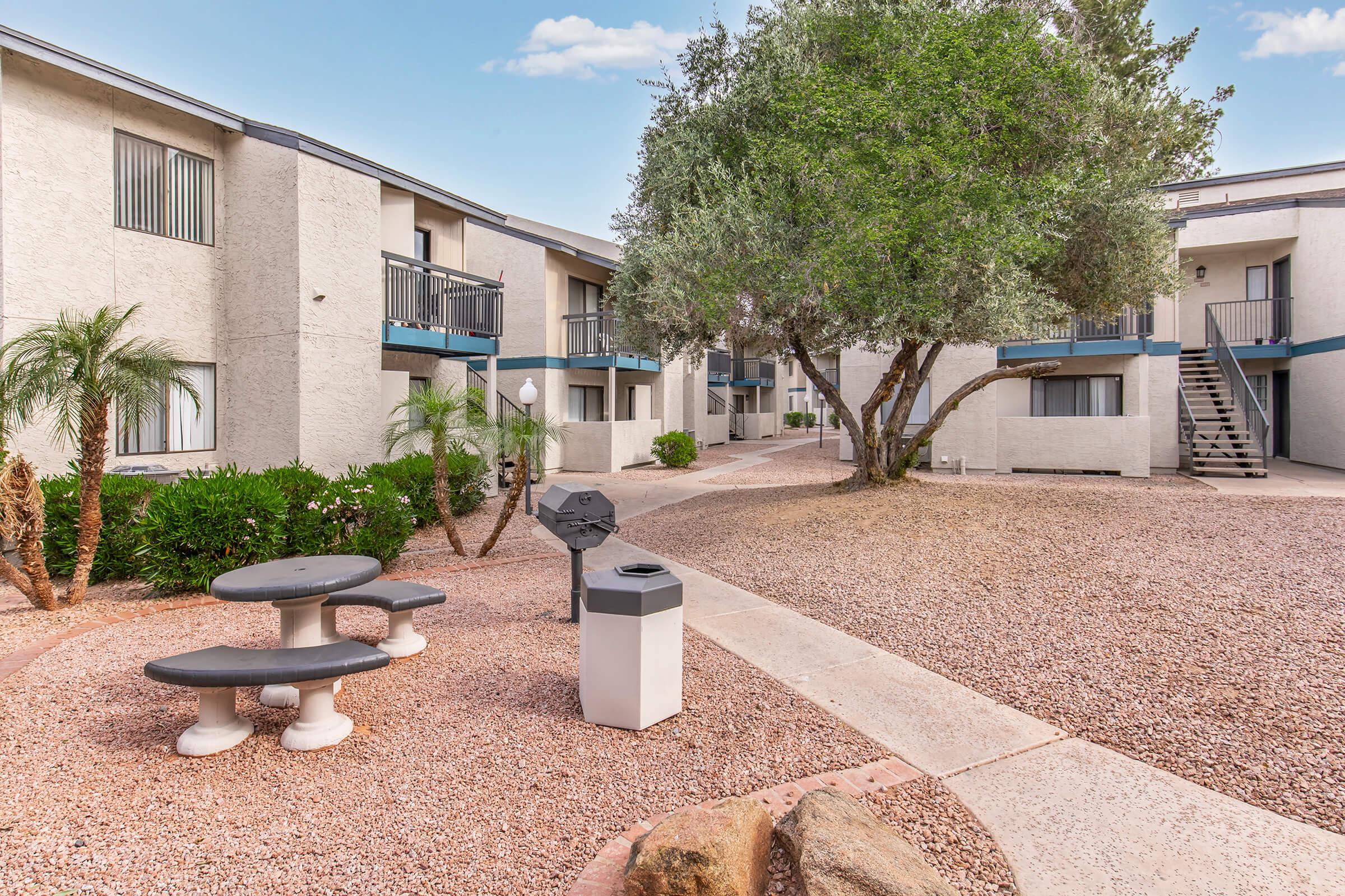 A courtyard area of an apartment complex featuring a picnic table, a trash bin, palm trees, and a well-maintained landscape with gravel paths. The buildings have multiple balconies and a staircase visible in the background, creating a welcoming outdoor space.