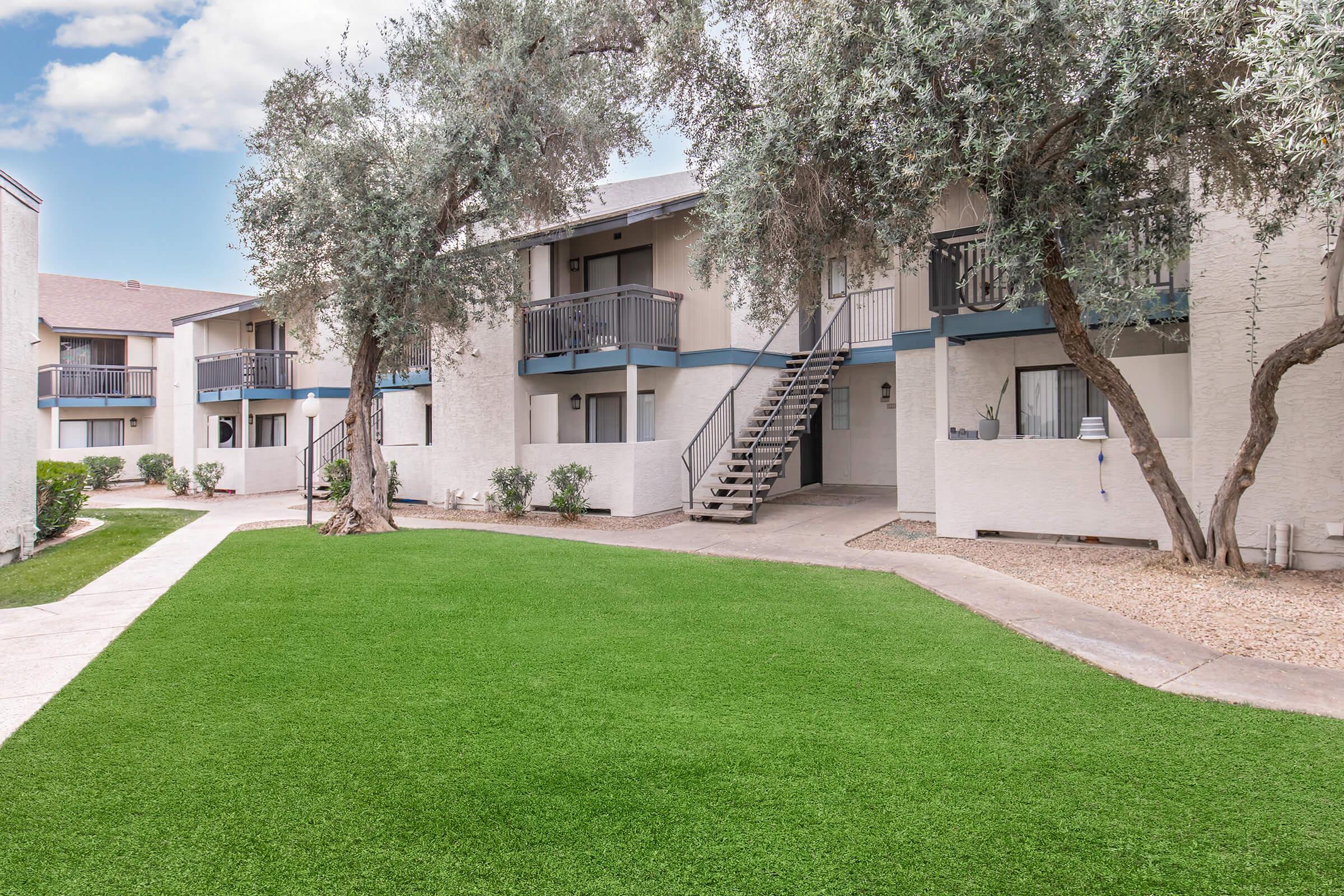 A well-maintained apartment complex featuring two-story buildings with balconies surrounded by landscaped green lawns and trees. A pathway runs through the area, leading to the entrance stairs of the units. Clear blue skies are visible in the background.