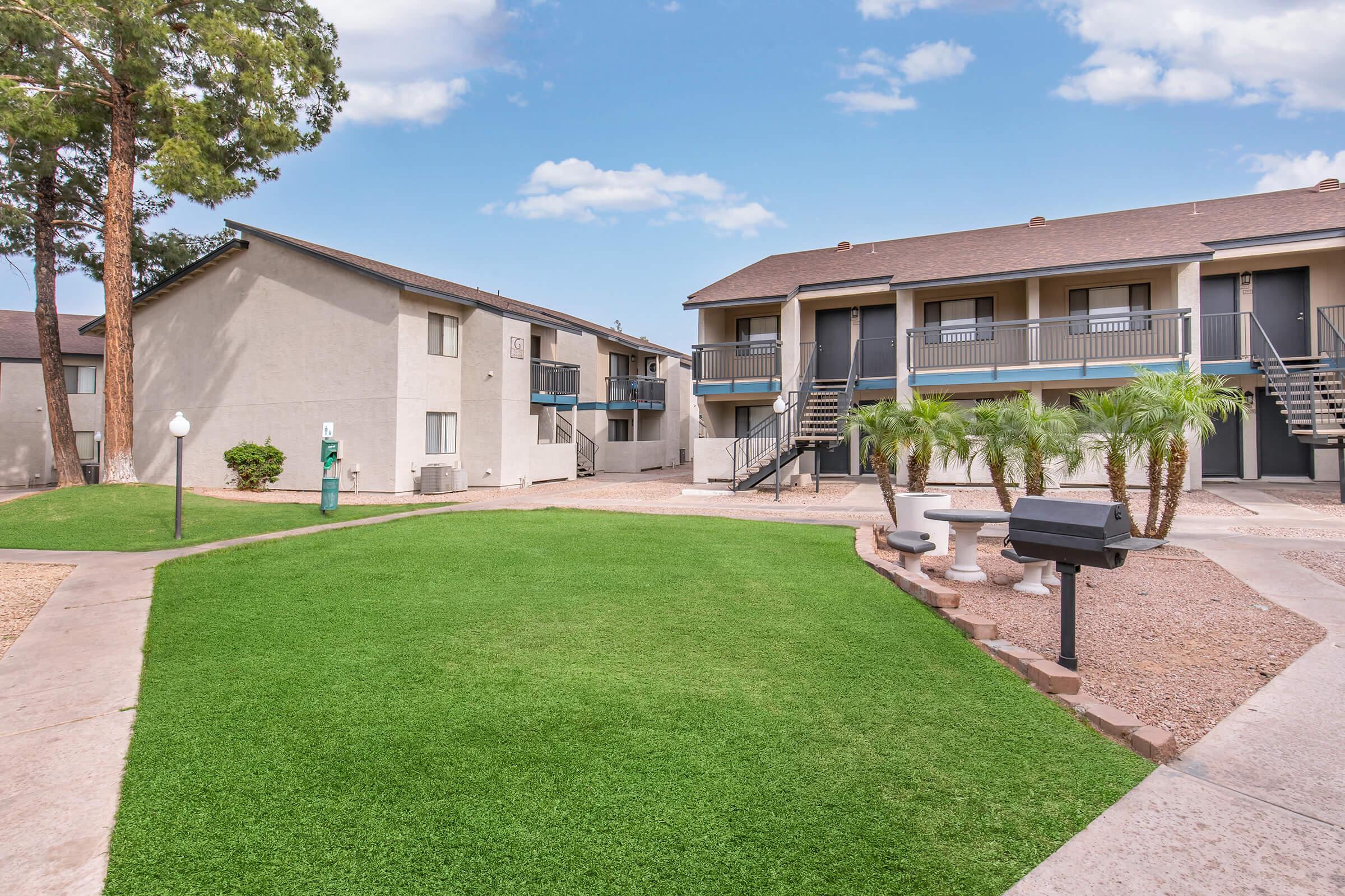 A view of a well-maintained apartment complex with two buildings surrounded by green grass. There are palm trees, a communal area with barbecue grills, and outdoor seating. Blue sky and scattered clouds are visible overhead, creating a pleasant atmosphere.