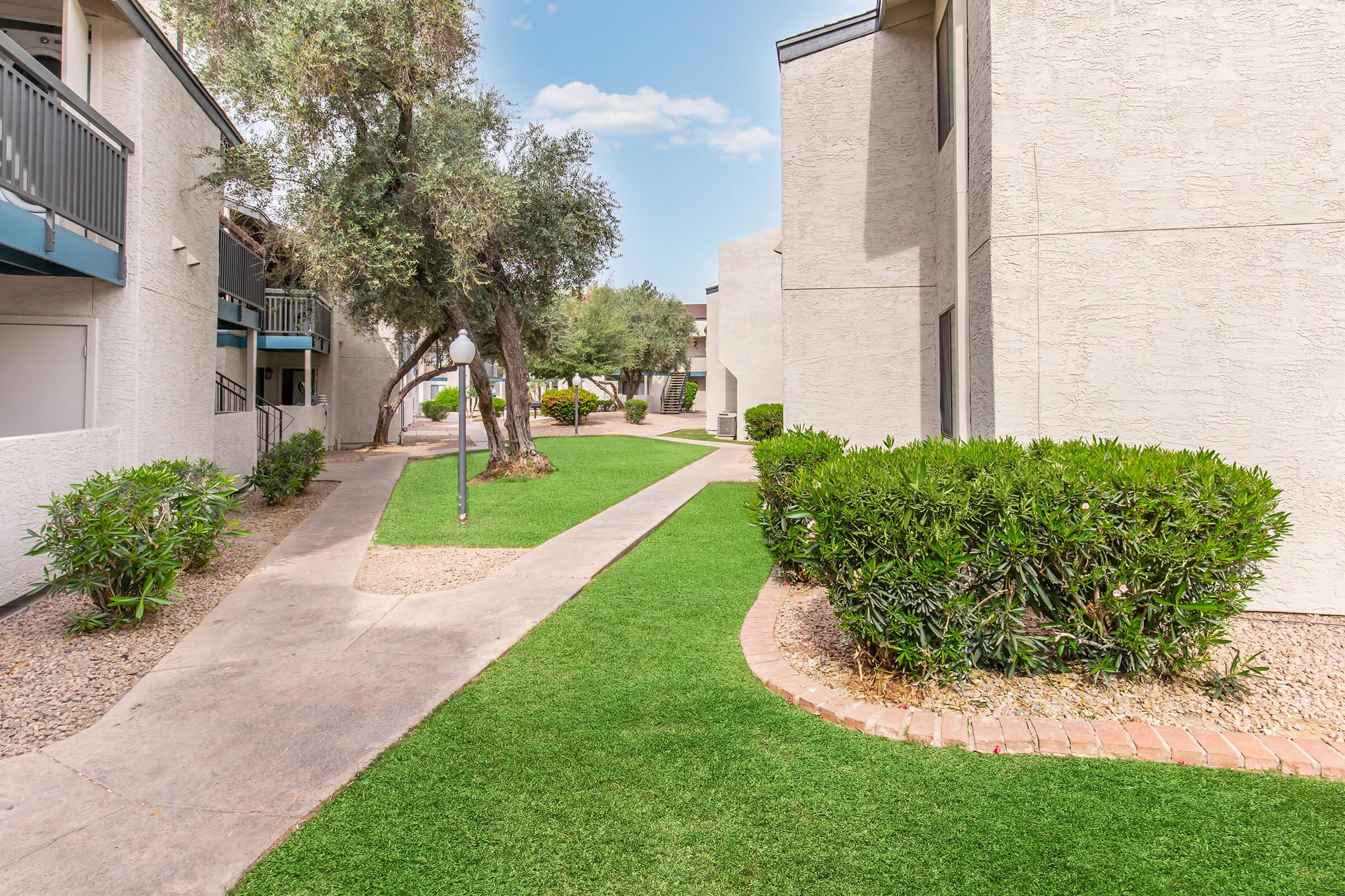 A landscaped pathway winds through a residential complex with green grass and shrubs. The buildings are beige, featuring balconies and a tree on the left side. The sky is blue with scattered clouds, creating a pleasant ambiance in the outdoor space.