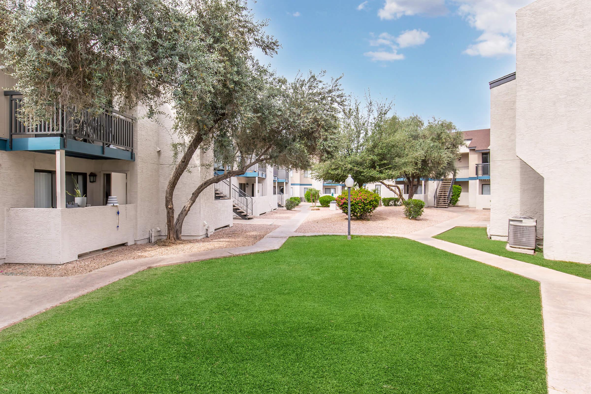 Landscape view of a well-maintained apartment complex with manicured green lawns and pathways. The scene includes several buildings, trees providing shade, and a bright blue sky with fluffy clouds. It's a serene and inviting environment conducive to outdoor living.