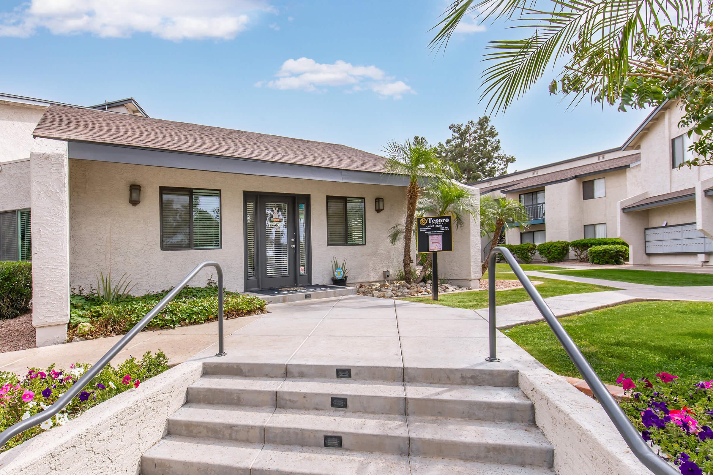 Exterior view of a residential complex featuring a modern office entrance with black-framed windows, a decorative sign, and steps leading up to the door. Lush green landscaping, flowering plants, and palm trees enhance the inviting atmosphere on a clear, sunny day.
