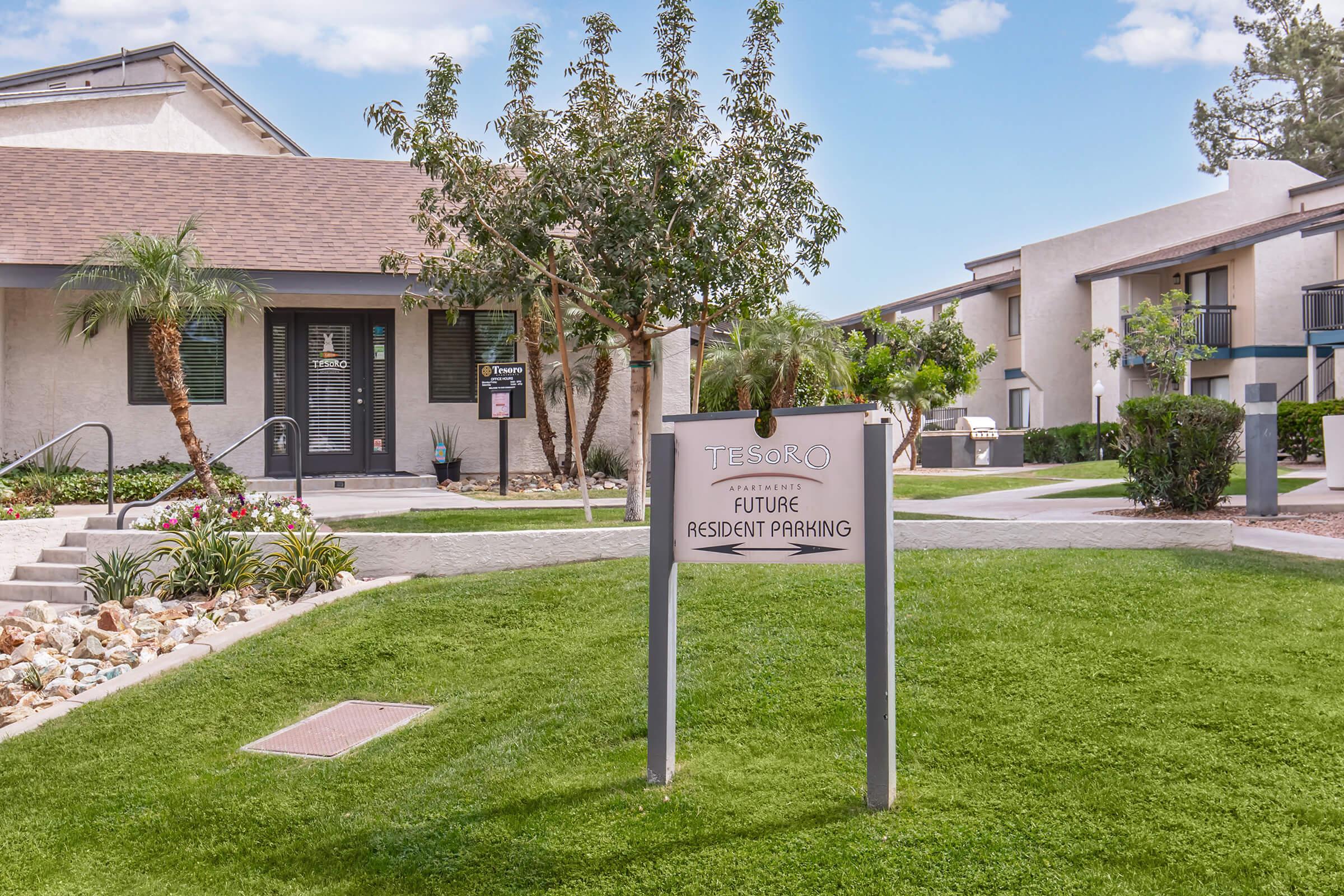 Sign for "Tesoro" indicating future resident parking, surrounded by well-maintained grass and landscaping. In the background, there are two residential buildings with balconies, and palm trees add a touch of greenery to the area. Clear blue sky overhead creates a pleasant atmosphere.