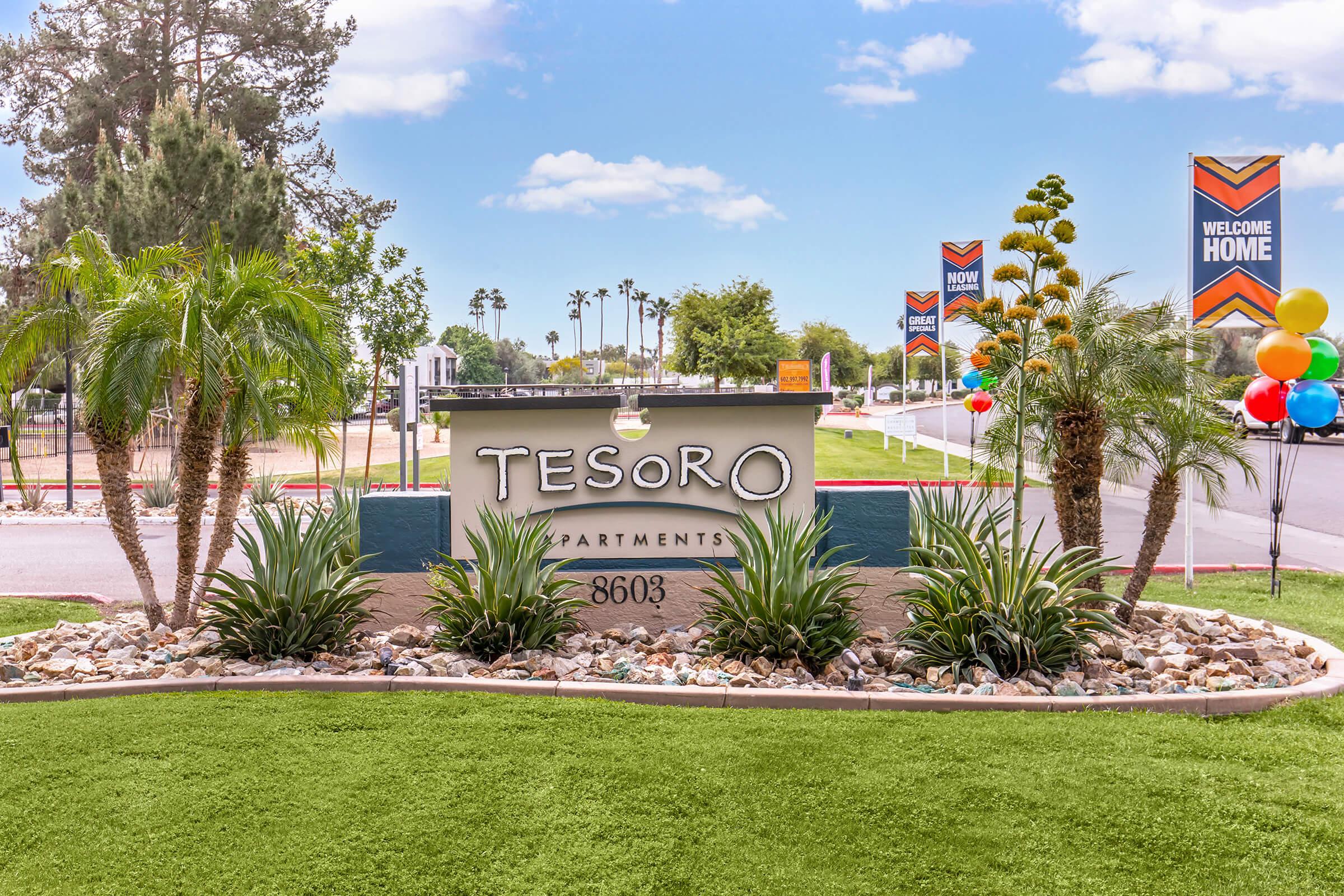 Sign for Tesoro Apartments featuring the name prominently displayed. The entryway includes decorative plants and stones, set against a backdrop of a clear blue sky. Colorful banners with "Welcome Home" are visible in the background, along with palm trees and festive balloons, creating a welcoming atmosphere.