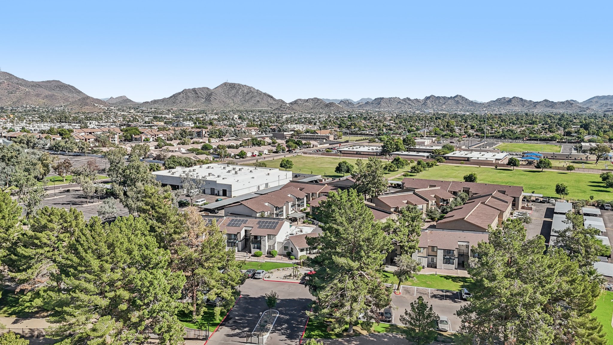 Aerial view of a suburban area featuring residential buildings, green spaces, and nearby mountains under a clear blue sky. The landscape includes trees and a mix of commercial and residential structures, illustrating a vibrant community with natural surroundings.