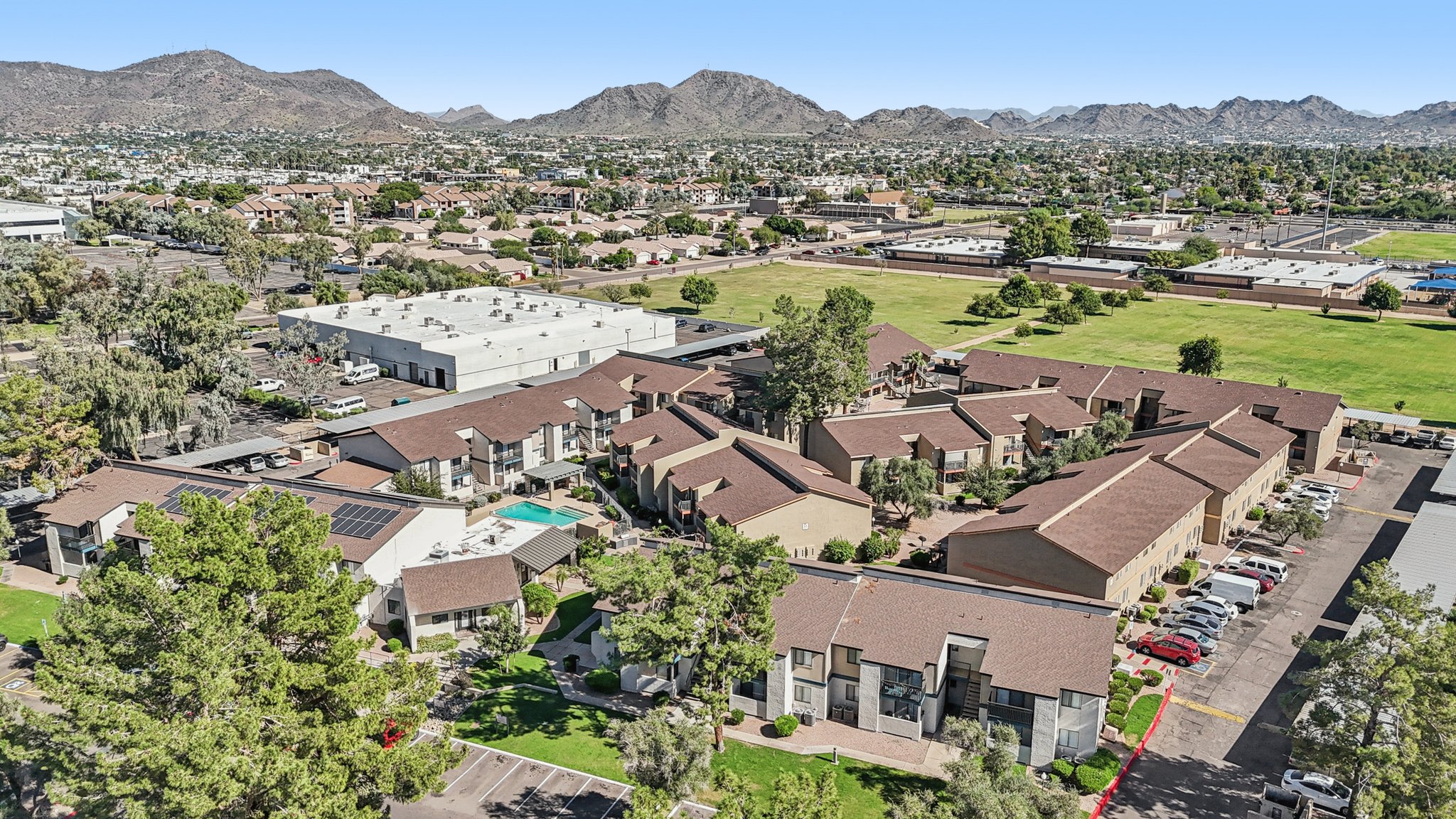 Aerial view of a residential area featuring several low-rise apartment buildings with brown roofs, a swimming pool, and surrounding green spaces. In the background, there are mountains and additional urban developments, with clear blue skies overhead.