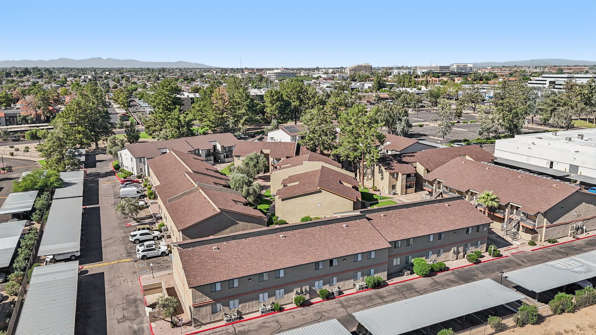 Aerial view of an apartment complex surrounded by trees, with buildings arranged in a U-shape. There are several parked cars along the driveways and a larger parking lot in the background. The landscape includes mountains in the distance under a clear blue sky.