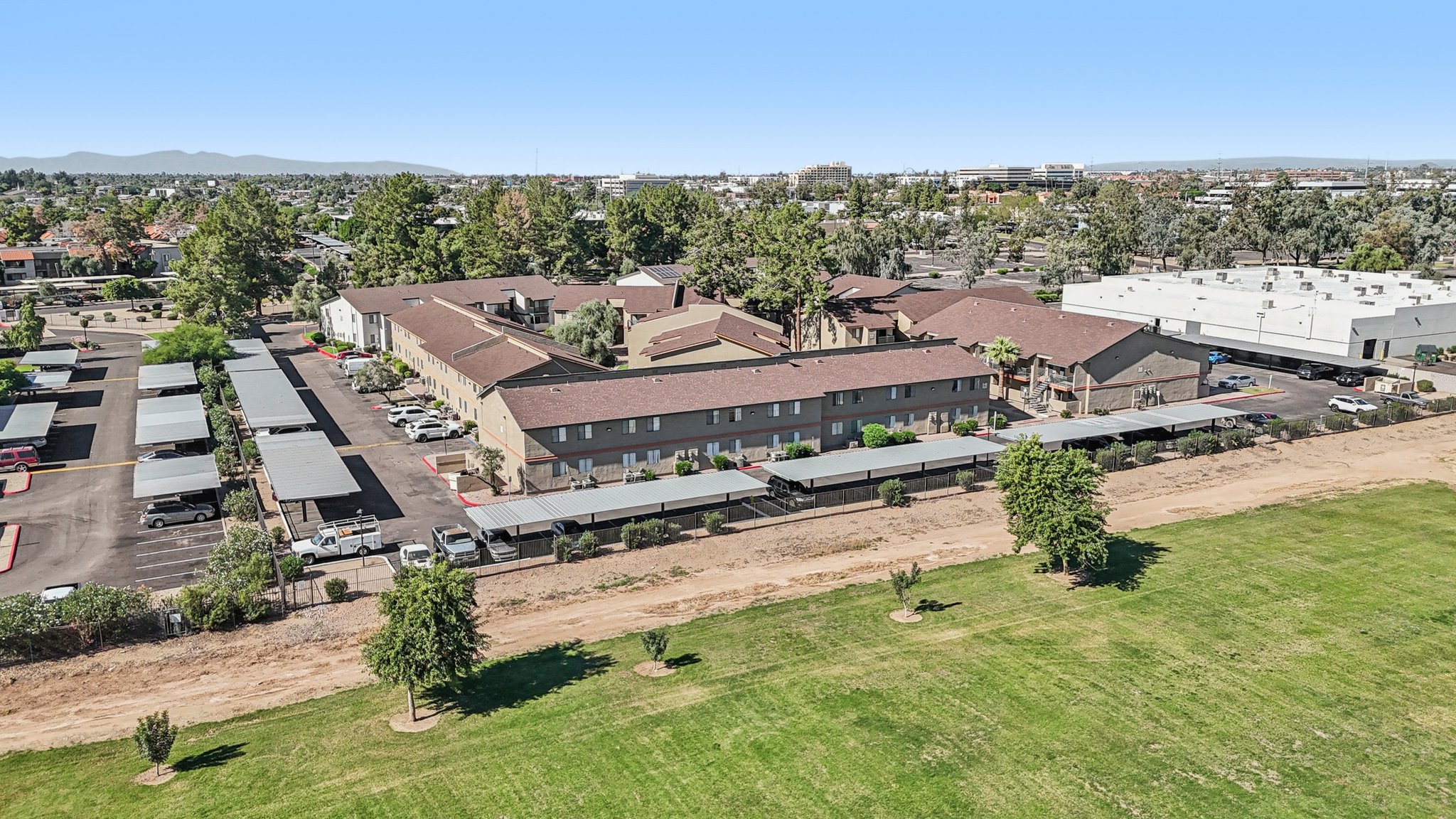 Aerial view of a multi-story building with a reddish-brown roof, surrounded by green grass and several parking spaces. Trees are visible along the perimeter, and there are additional buildings in the background. The area appears to be suburban, with clear skies above.