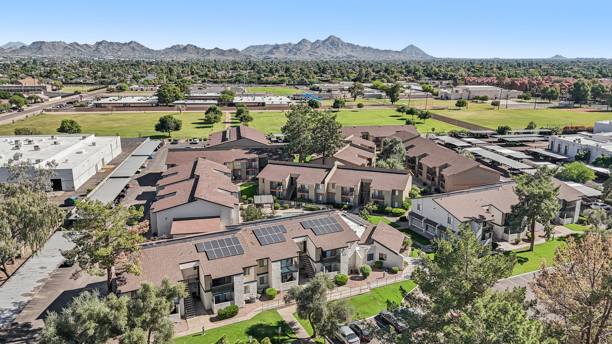 Aerial view of a residential complex with several buildings featuring brown roofs. Solar panels are visible on some roofs. In the background, a mountain range is situated against a clear blue sky, while green fields and additional structures can be seen nearby.