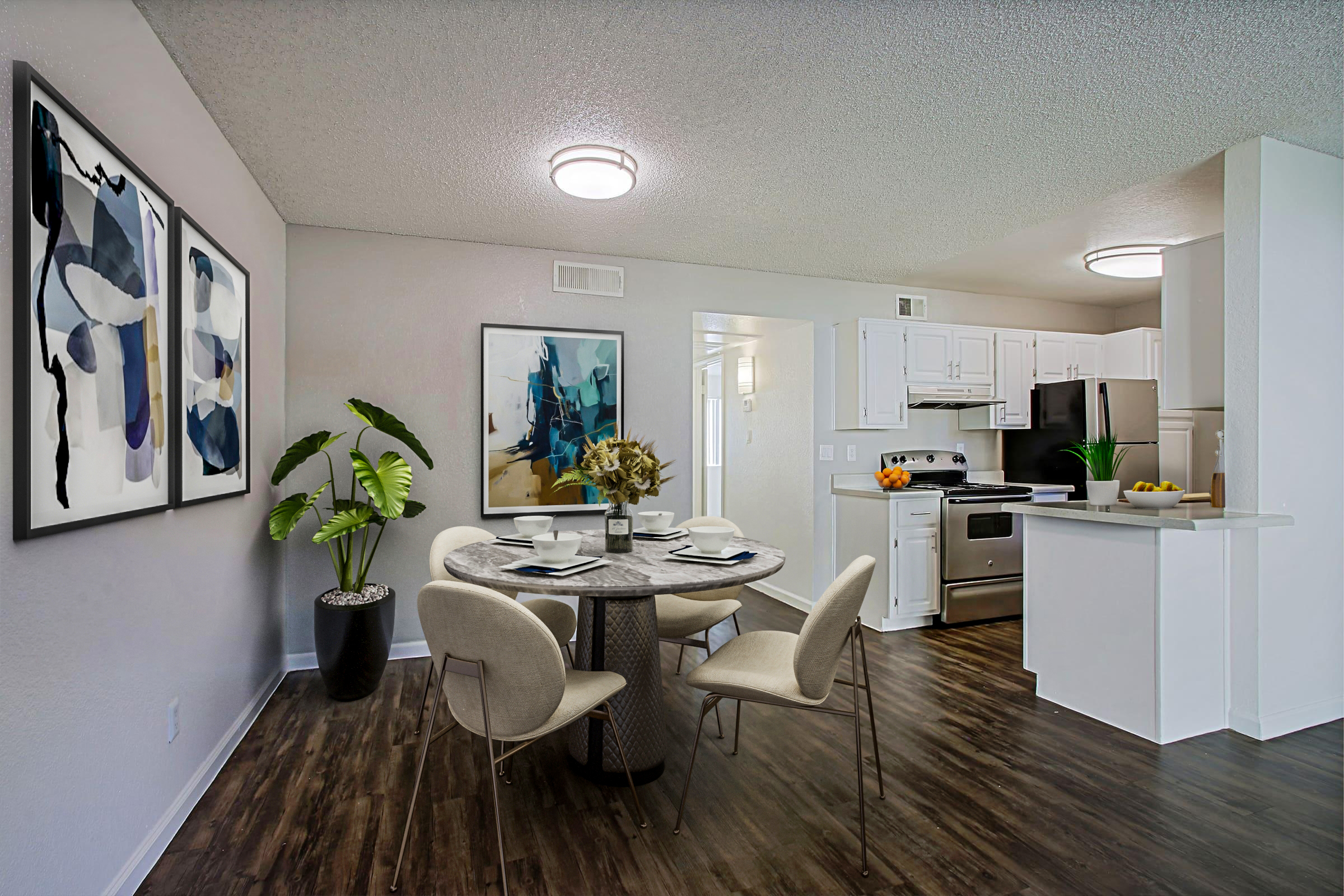 Modern apartment interior featuring a dining area with a round table and four chairs, a kitchen with stainless steel appliances, and contemporary art on the walls. A large plant adds a touch of greenery, while the space is brightly lit with ceiling lights. The flooring is dark wood, creating a warm atmosphere.