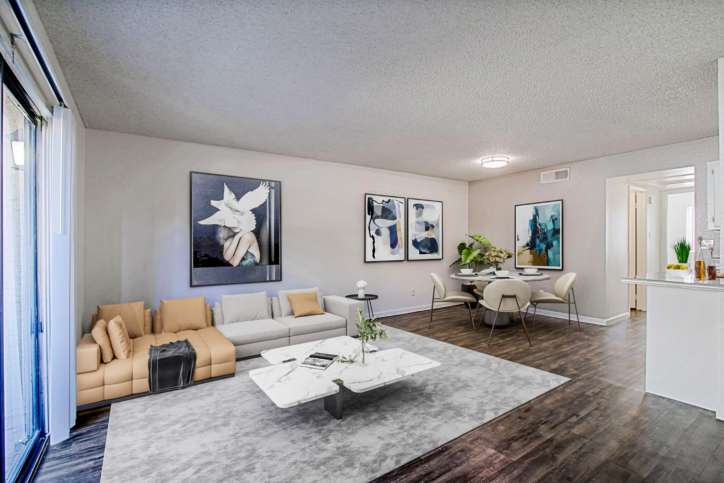 A modern living room featuring a beige sectional sofa with decorative cushions, a marble coffee table, and a large area rug. The walls are adorned with abstract artwork. A dining area is visible, with a round table and chairs. Natural light fills the space through a sliding glass door. The overall decor is minimalist and elegant.
